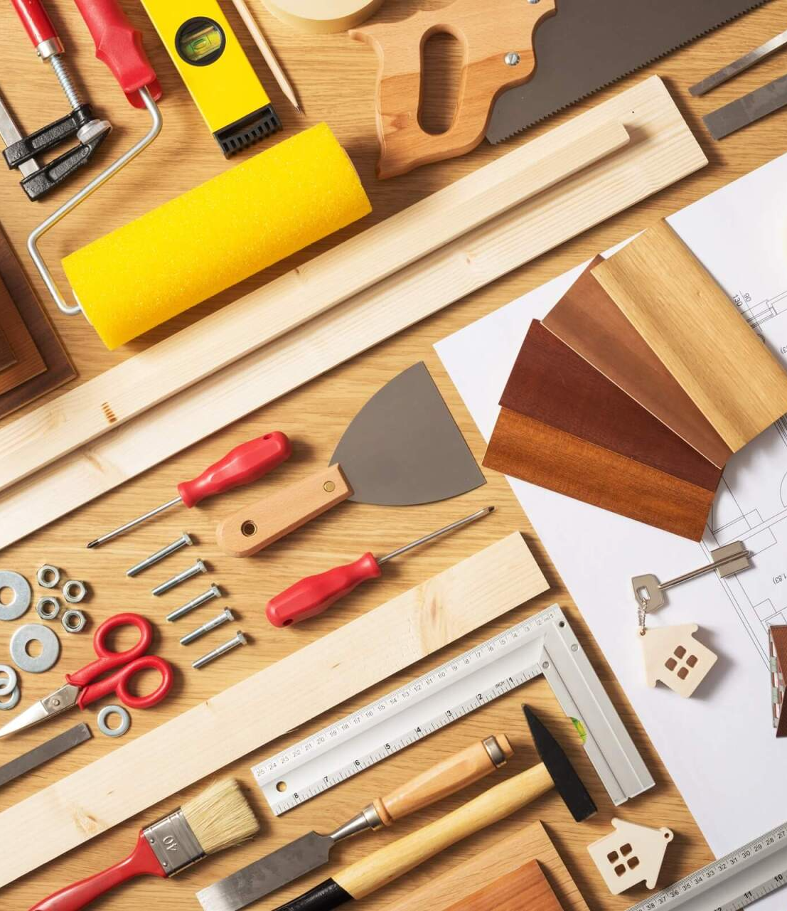 Assortment of woodworking tools and materials on a wooden surface: paint roller, saw, screws, wood samples, and blueprints.