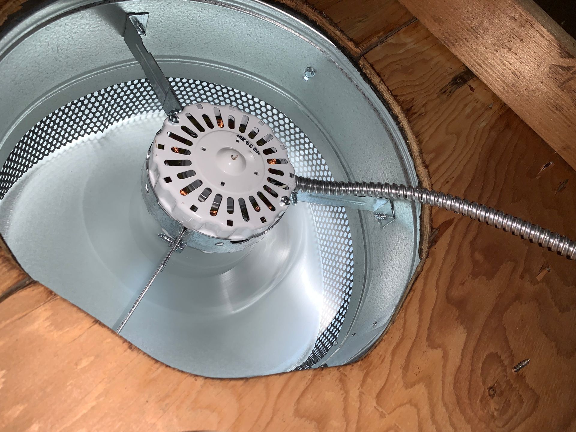 Motor inside a round, perforated metal duct, mounted in a wooden frame. Flexible conduit extends from the motor.