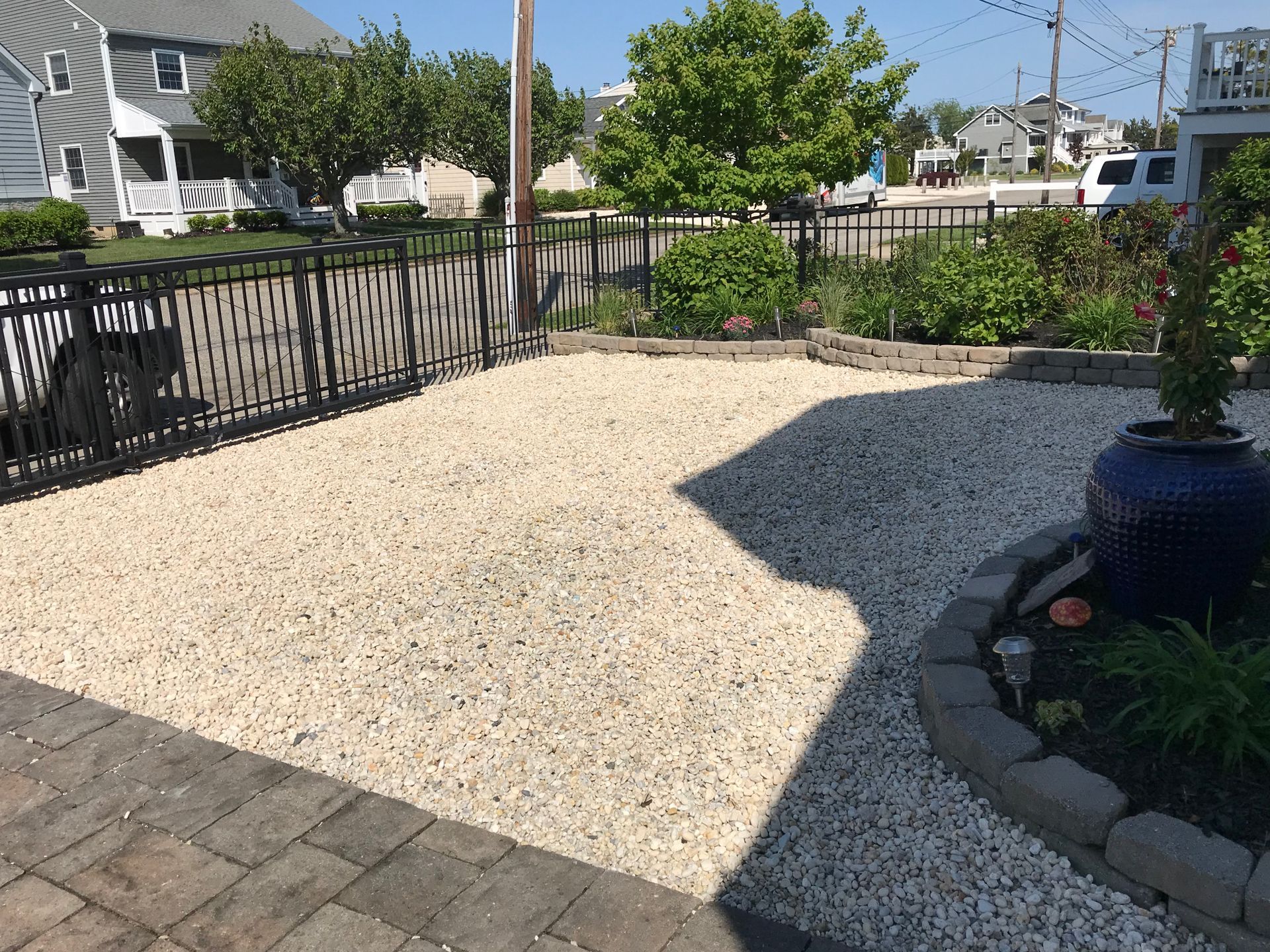 Gravel driveway with black metal fence, bordering flower beds and homes on a sunny day.
