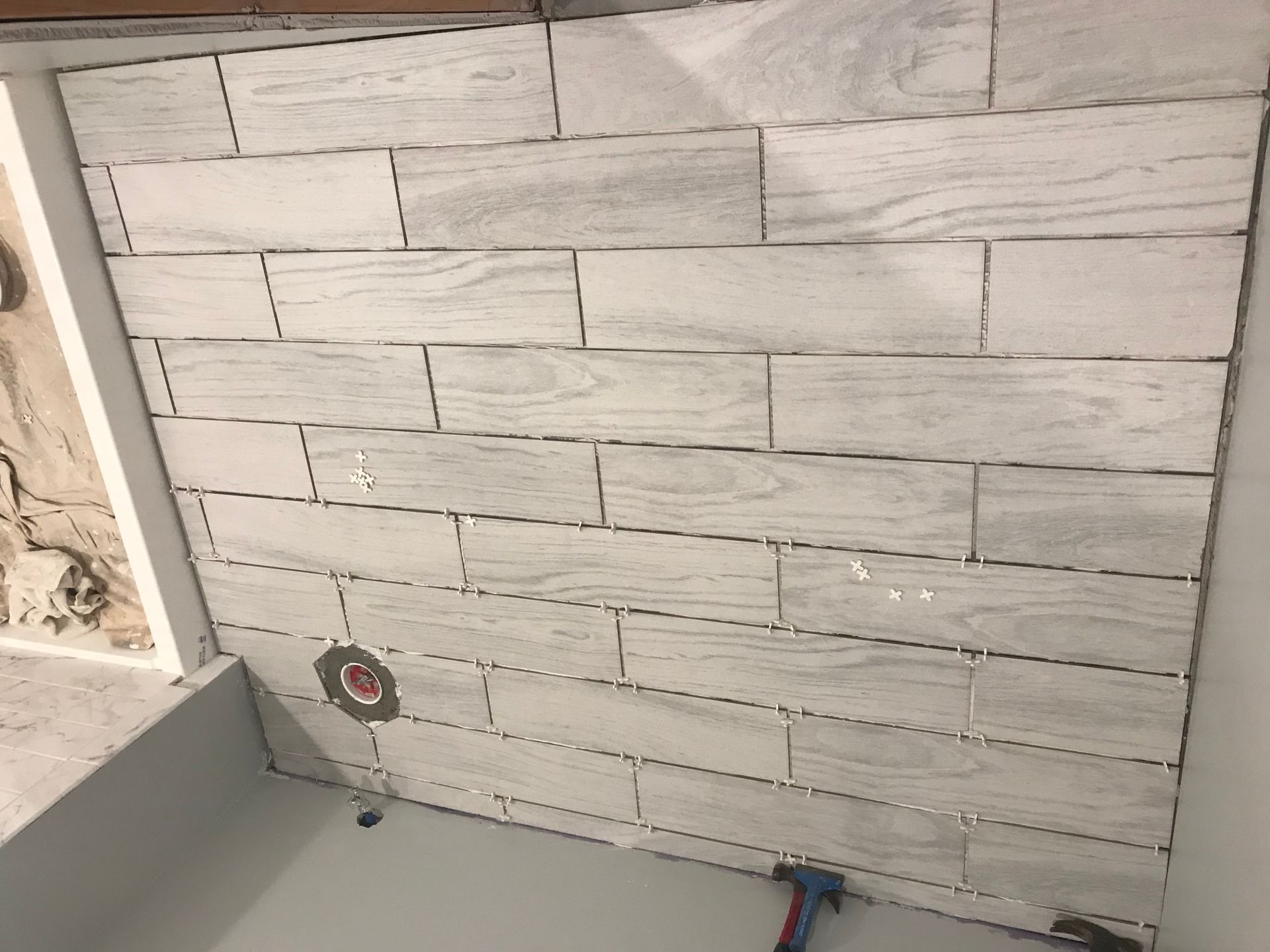 Tiled wall with light gray rectangular tiles, grout lines visible. Plumbing fixture and tools present.