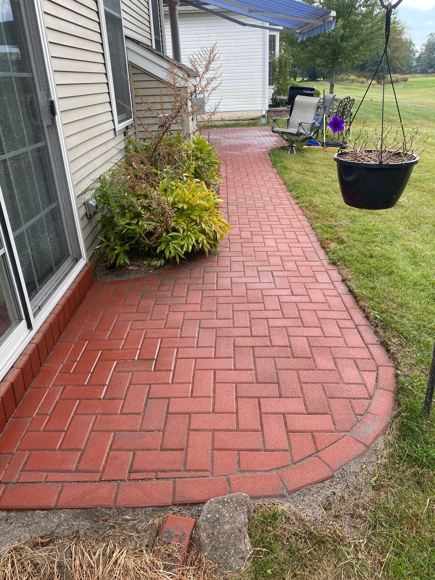 Red brick patio path alongside a beige house, with greenery and a hanging plant.