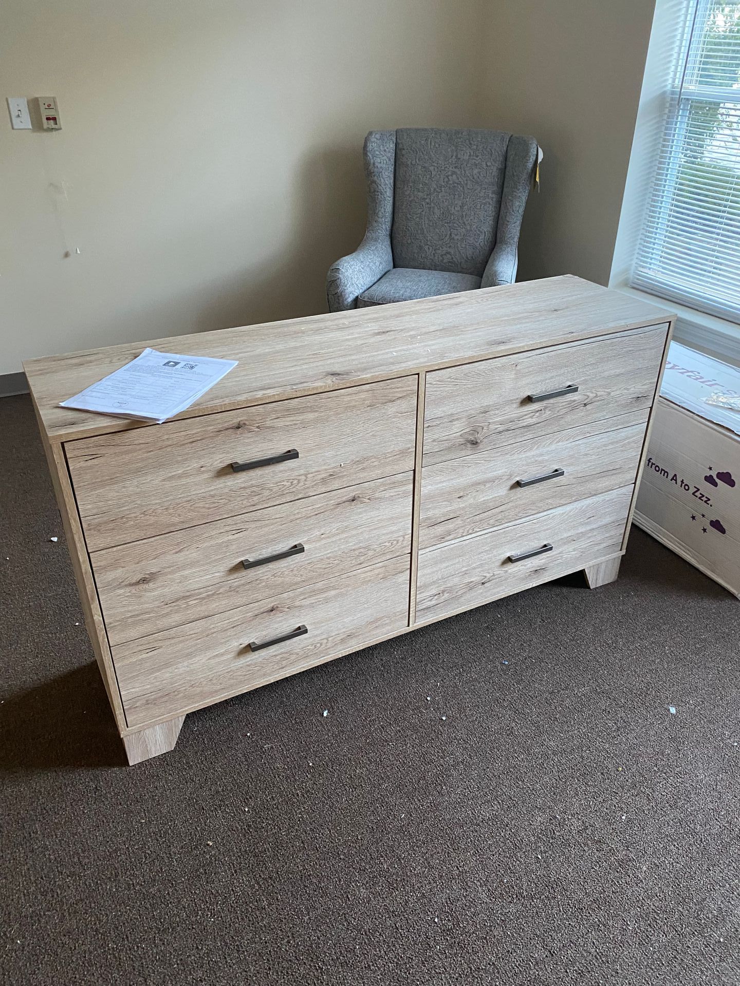 Light wood dresser with six drawers, gray wingback chair, and window in the background.