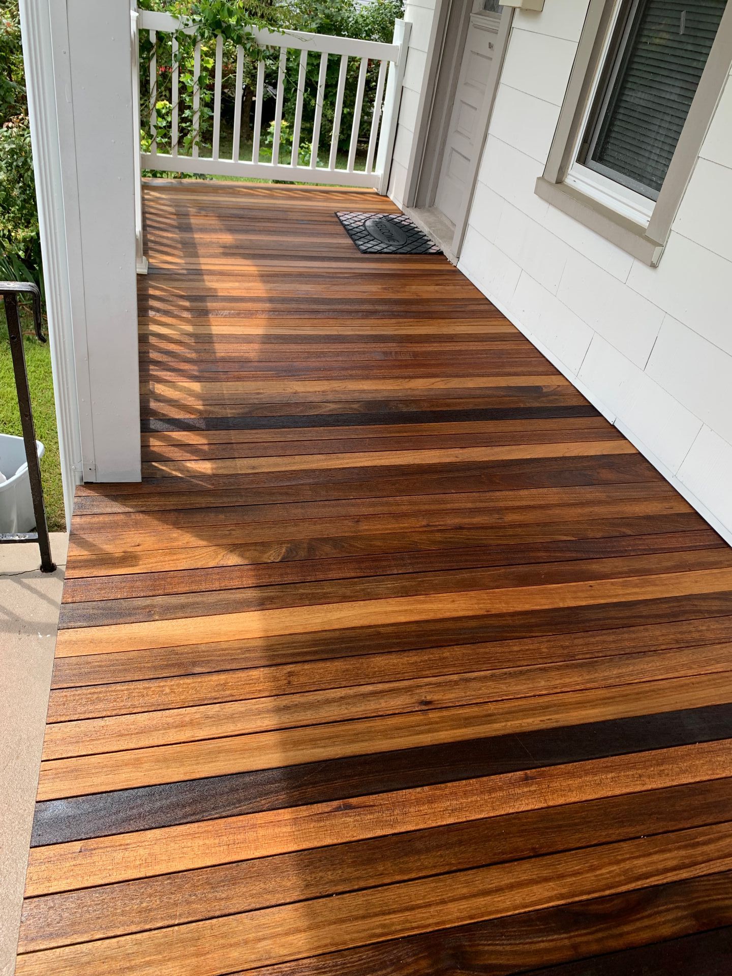 Wooden deck with dark and light brown stripes, outside a white building with porch and railing.