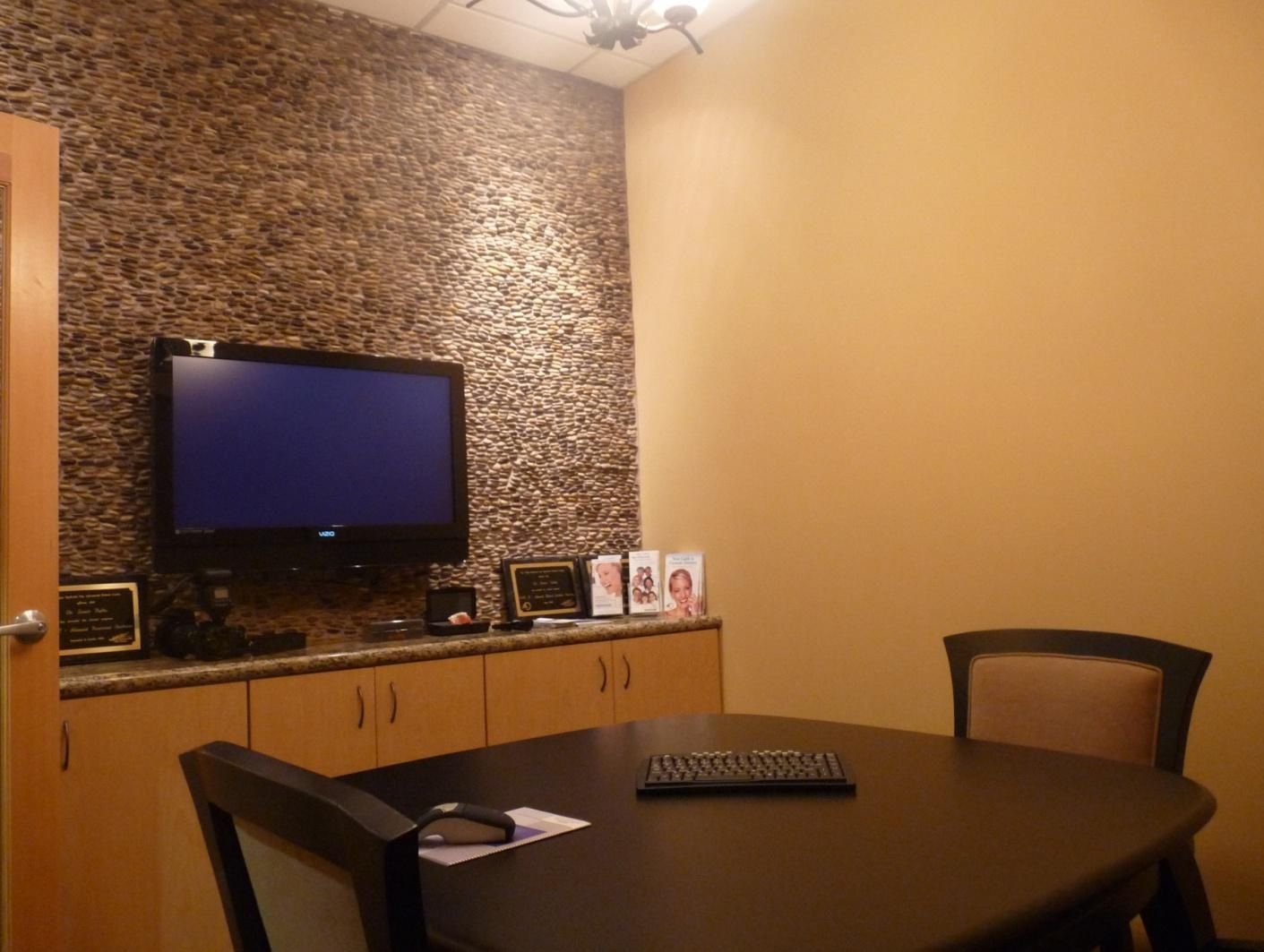 A room with a TV, table, and two chairs. A textured wall, and wooden cabinets.