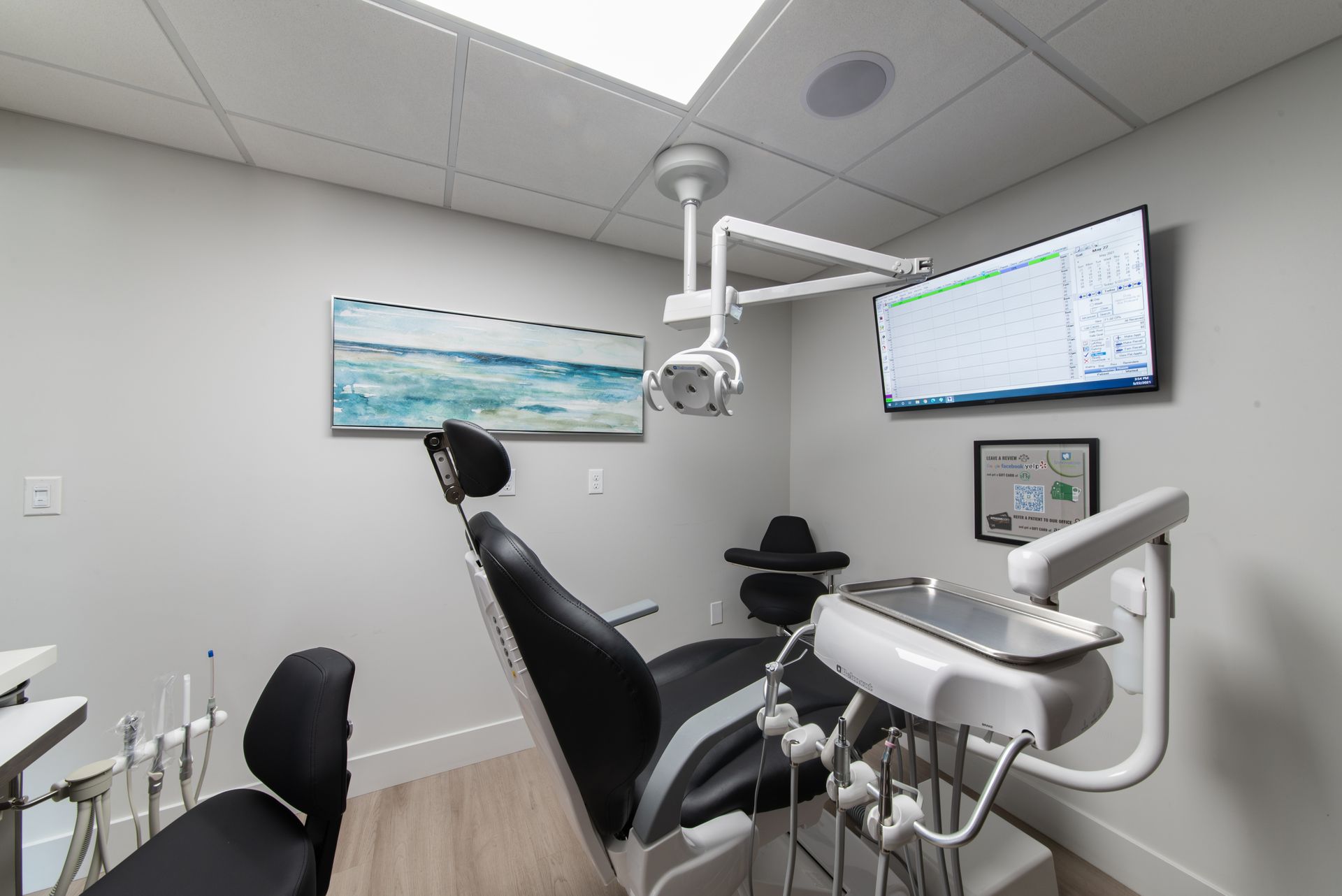 Dental exam room with dental chair, tools, and monitor. Neutral colors, bright overhead light.