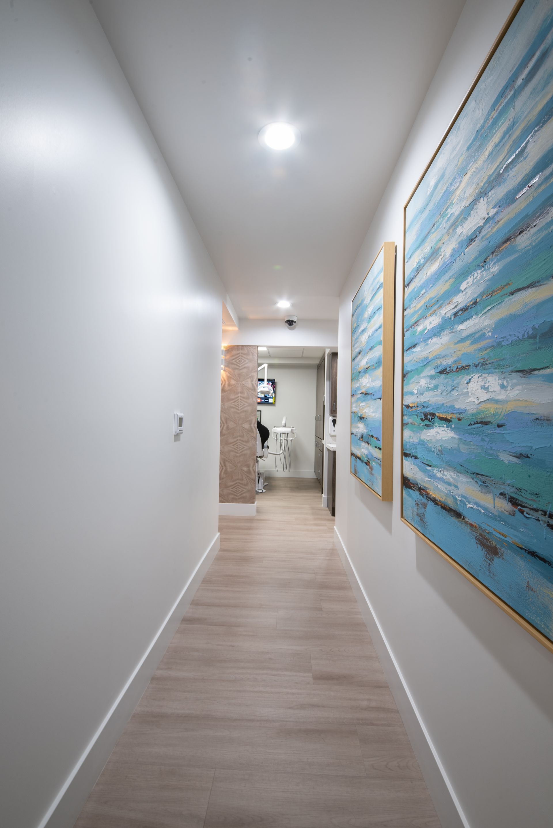 Long hallway in dental office, light wood floors, white walls, blue artwork.