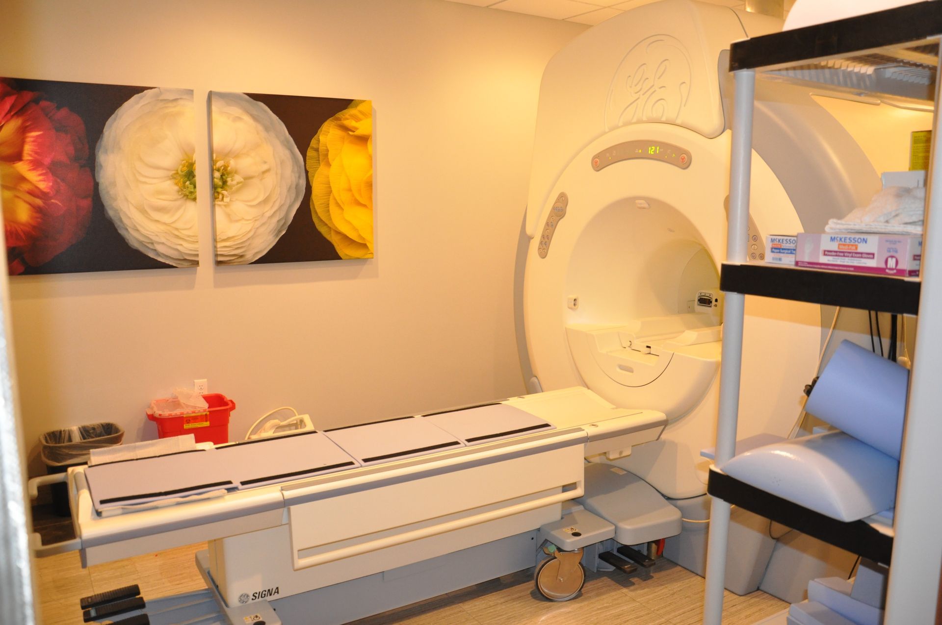 An MRI machine in a medical room, with a patient table, storage shelves, and colorful floral art on the wall.
