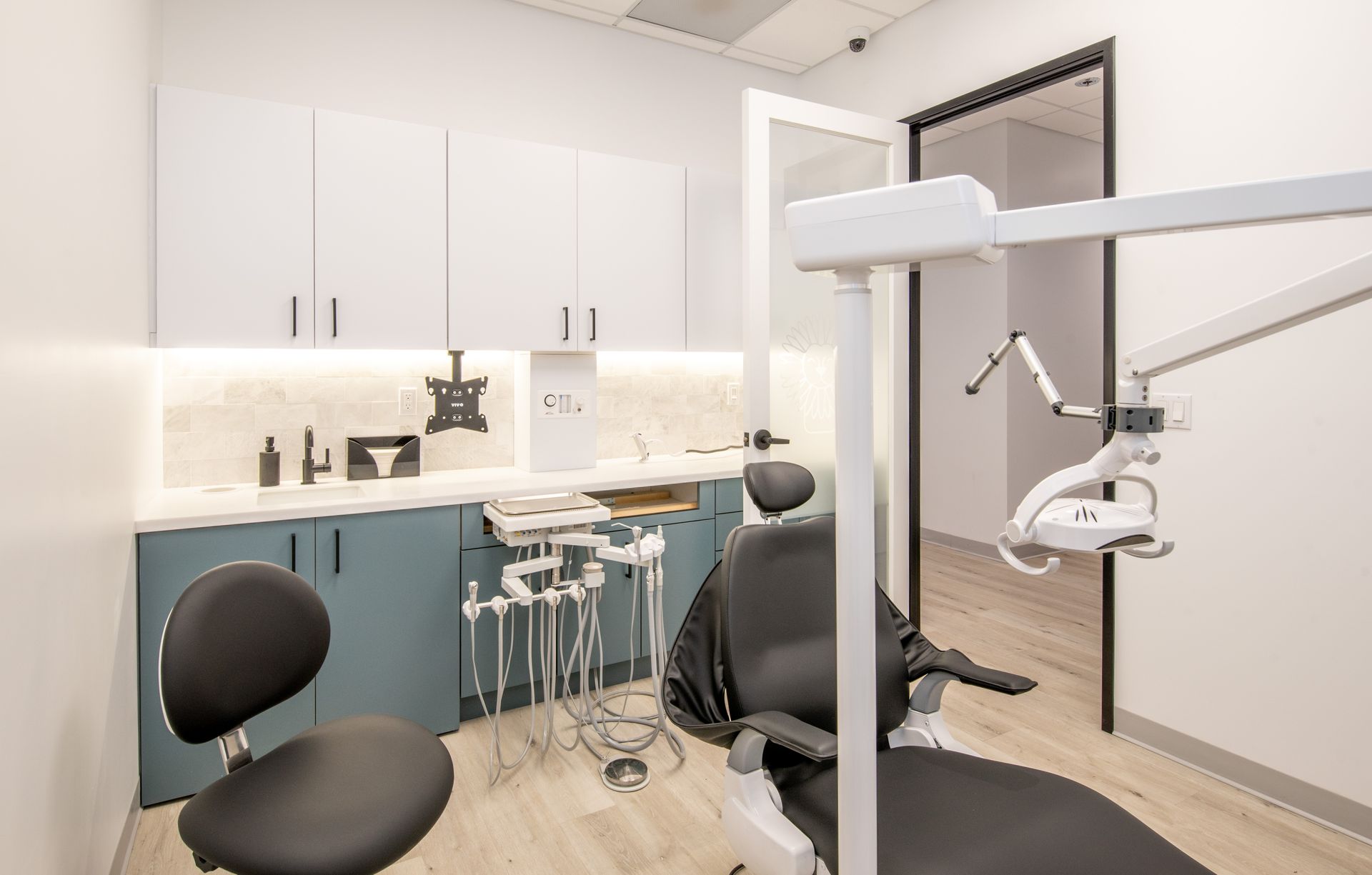 Dental operatory with chair, equipment, white and blue cabinetry, and an open door.