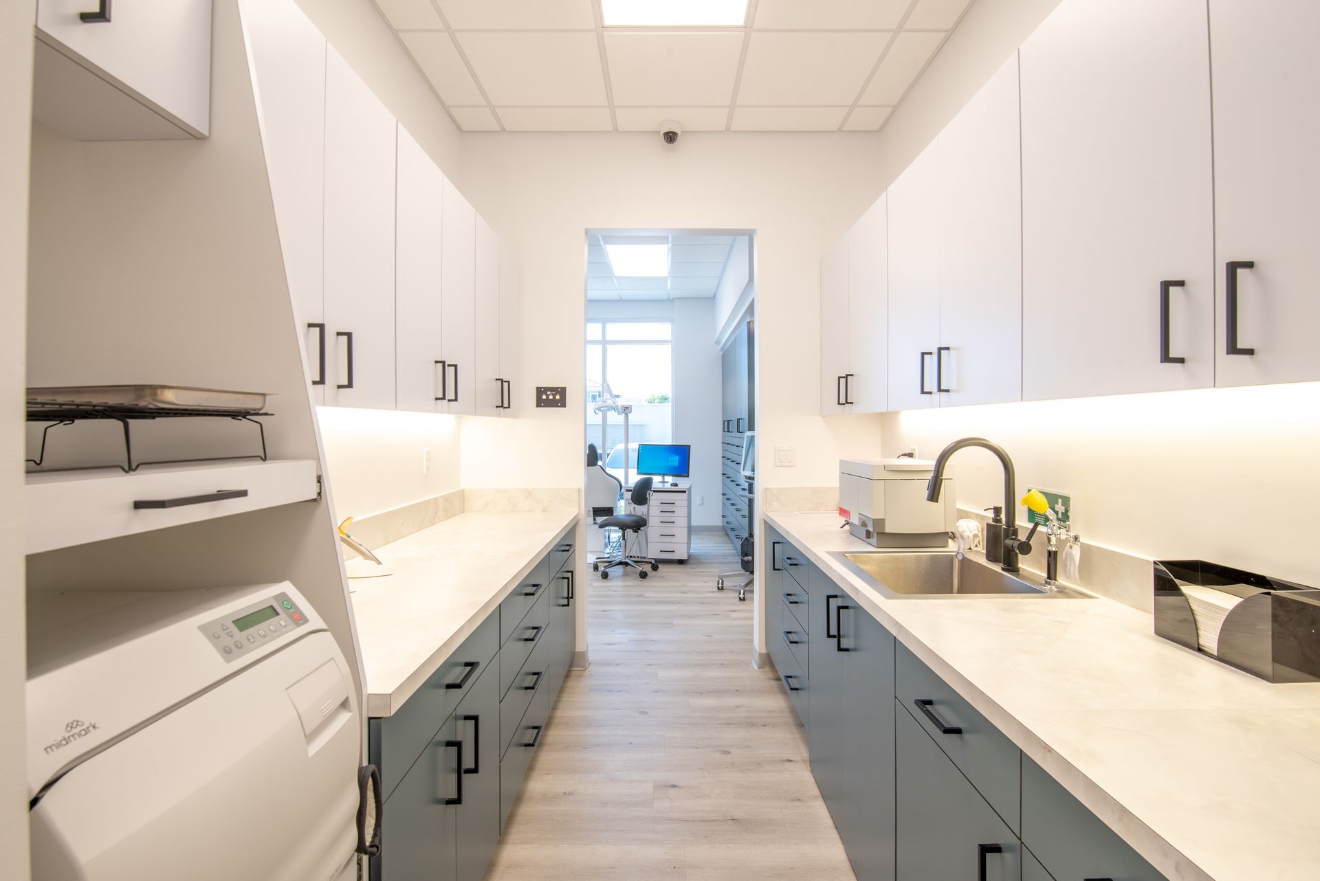 Dental office sterilization room, long, white cabinets, blue drawers, sink, sterilizer, open doorway to another room with desk.