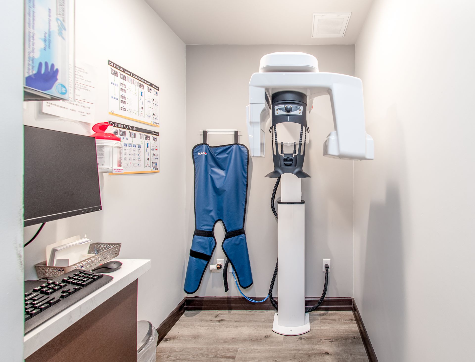 A dental office with an x-ray machine, computer, protective apron, and a white and grey color scheme.