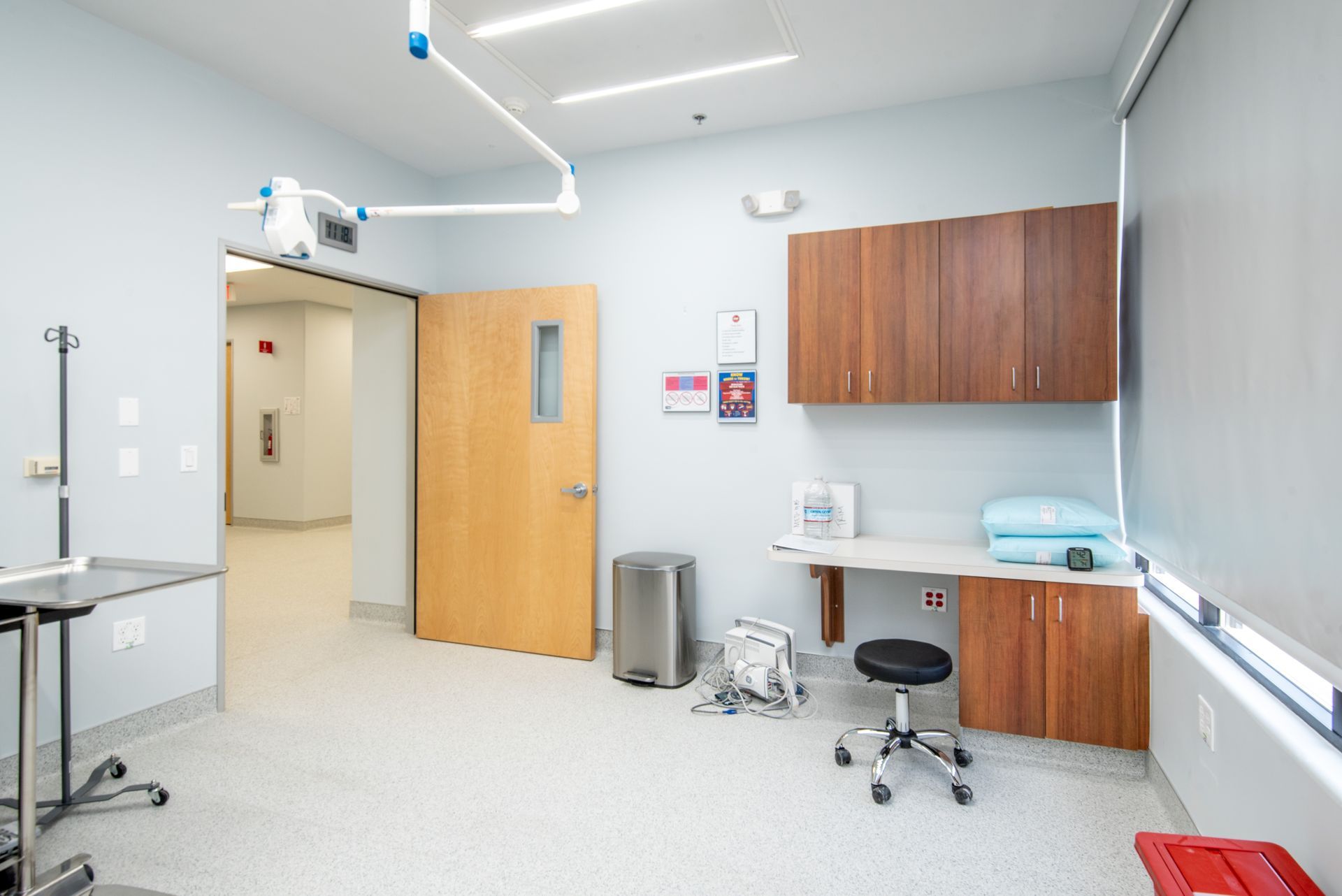 An empty examination room with light blue walls and wooden cabinets, with a desk, stool, and equipment.