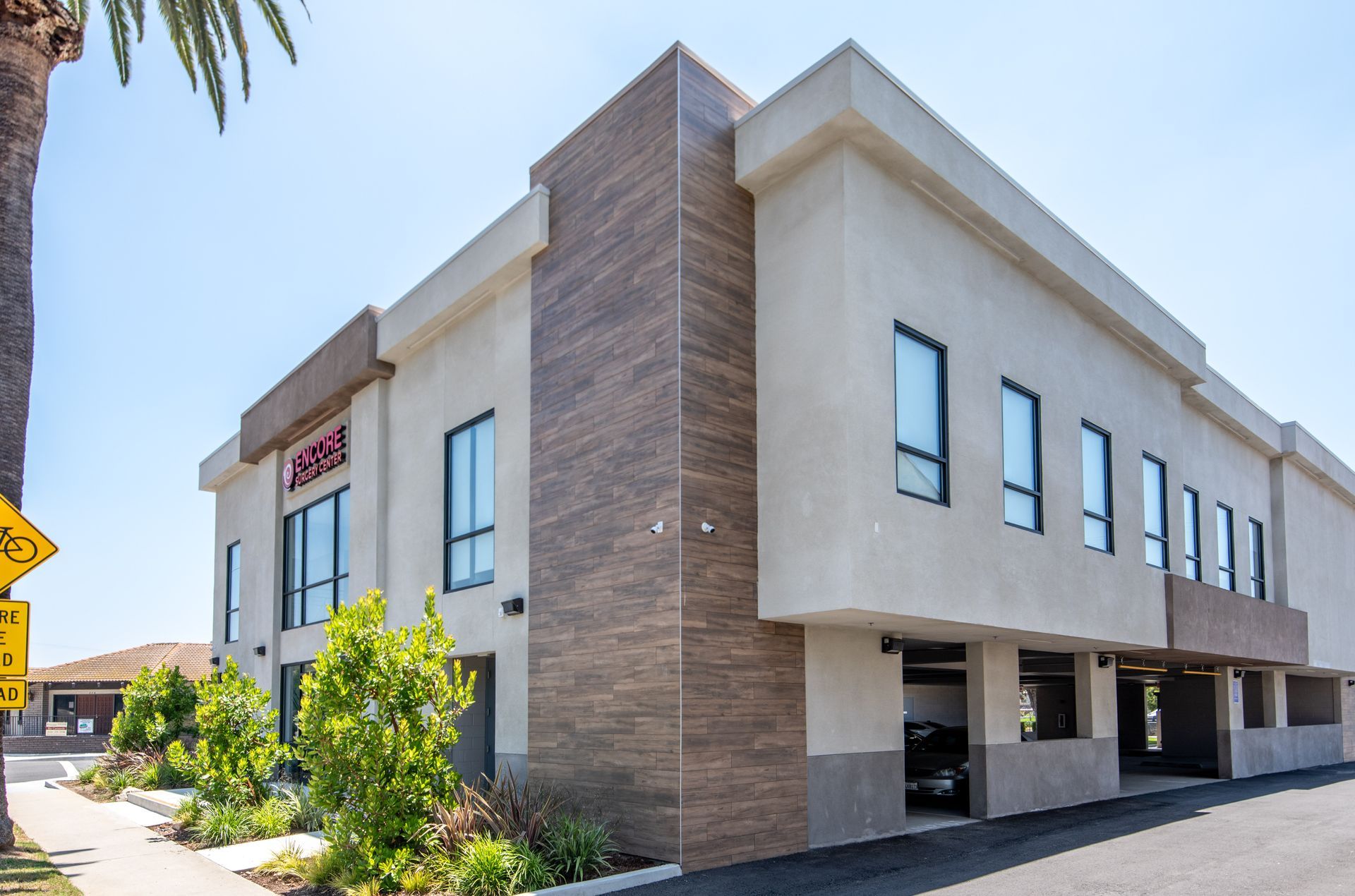 Two-story beige building with brown paneling, small windows, and parking underneath; sunny day.