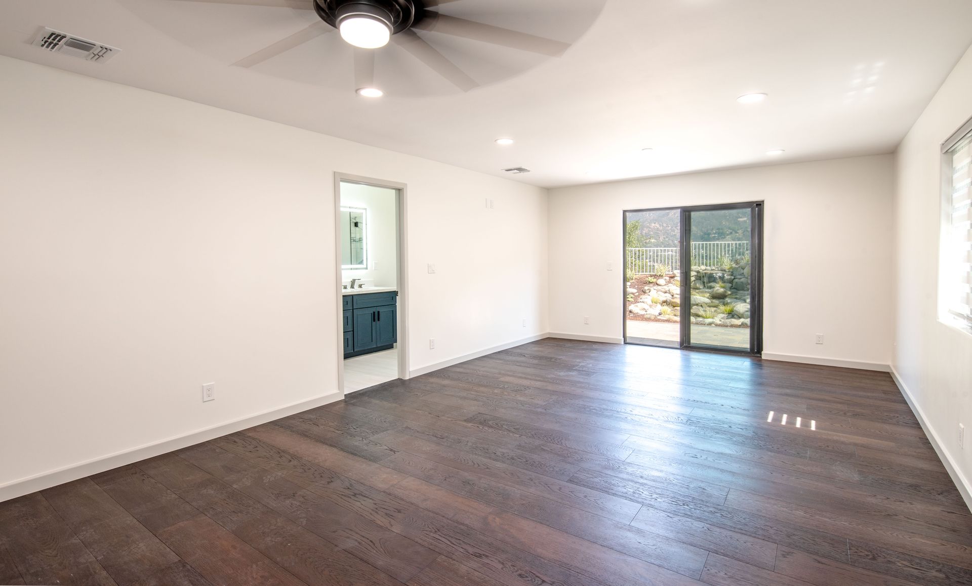Empty bedroom with dark hardwood floors, white walls, sliding glass door to backyard.