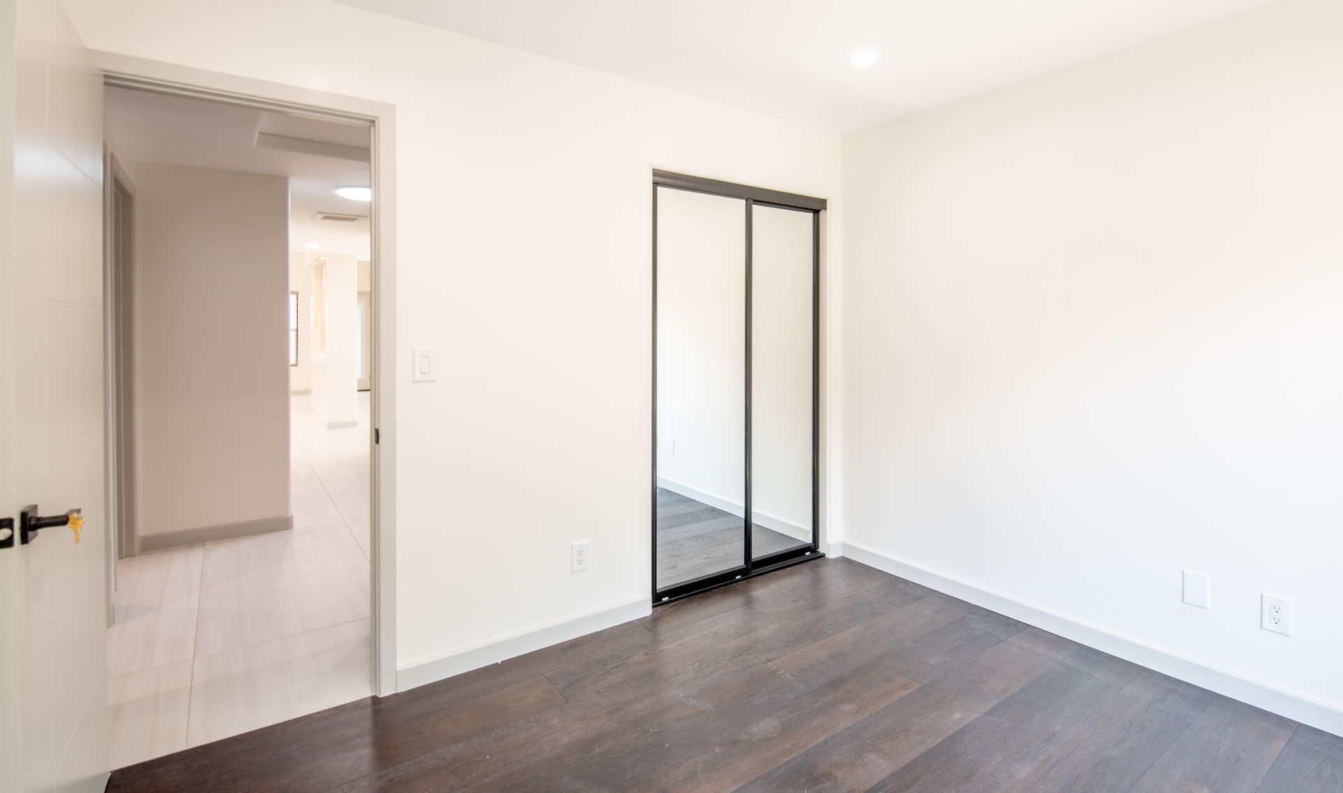 Empty bedroom with dark wood floors, white walls, sliding mirrored closet doors, and open doorway.