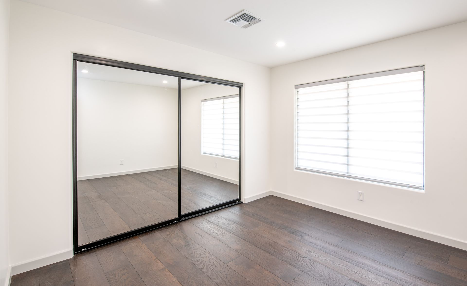 Empty bedroom with mirrored closet, window, and wood floors.