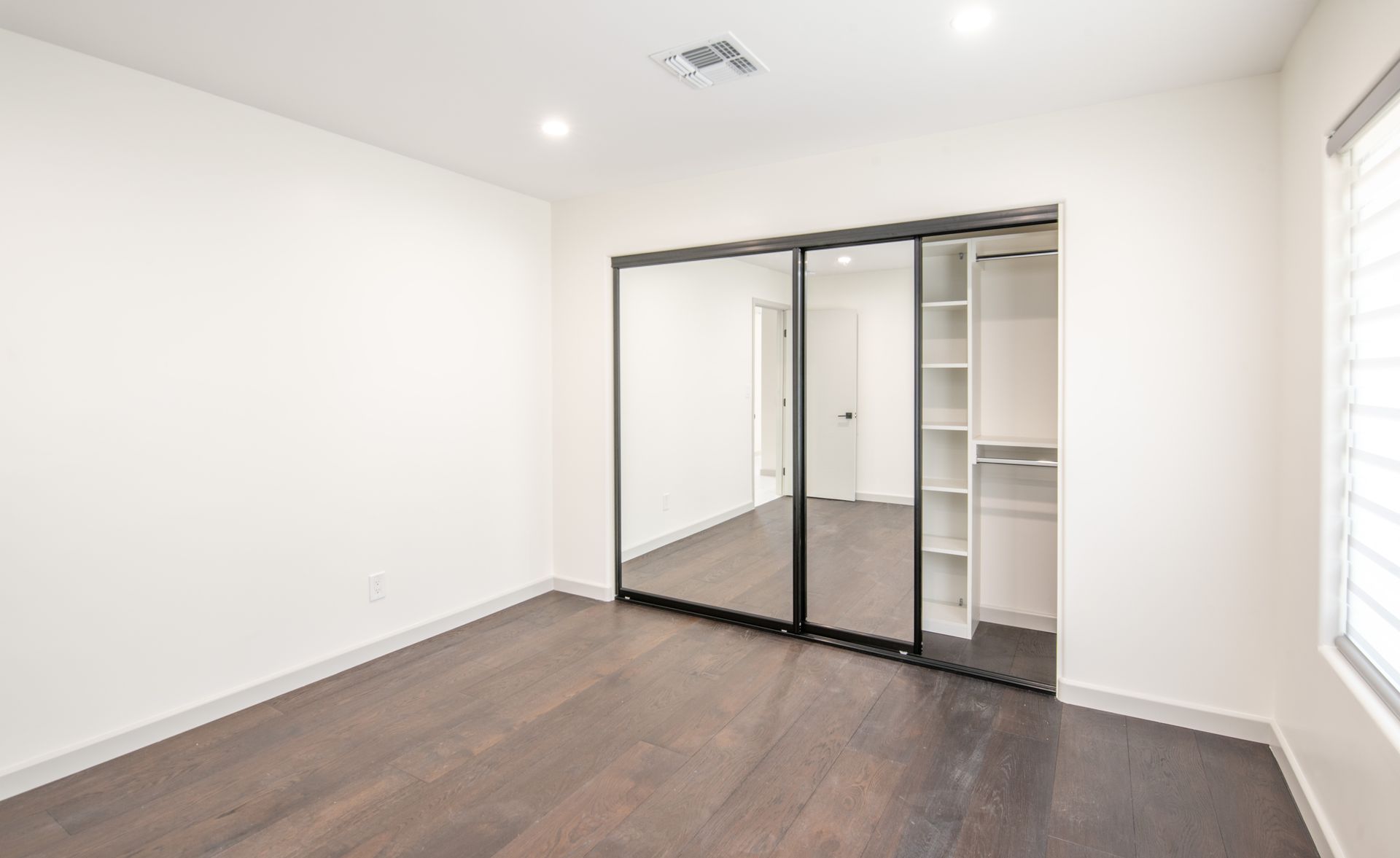 Empty bedroom with dark wood floors, mirrored closet, and built-in shelves.