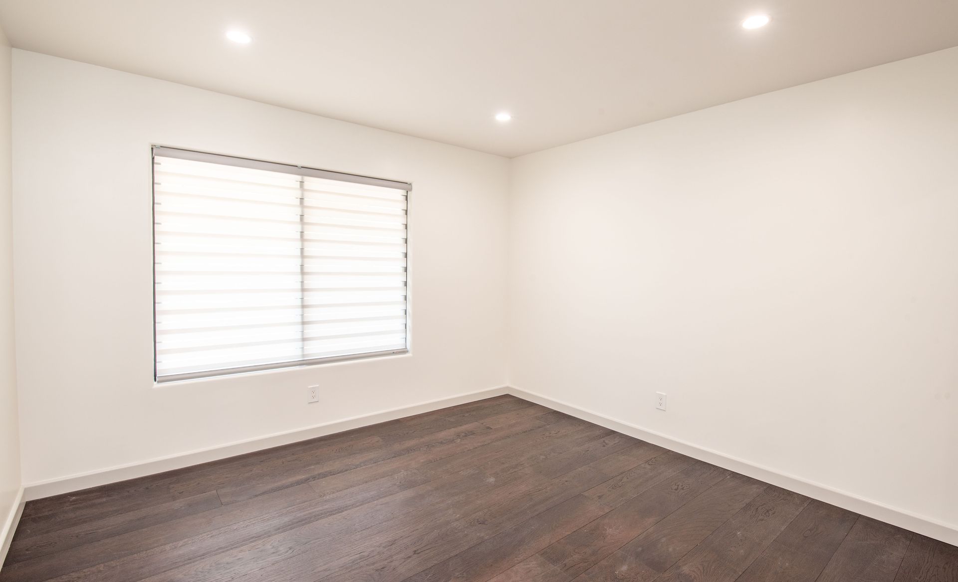 Empty room with dark wood floors, white walls, window with blinds, and recessed lighting.