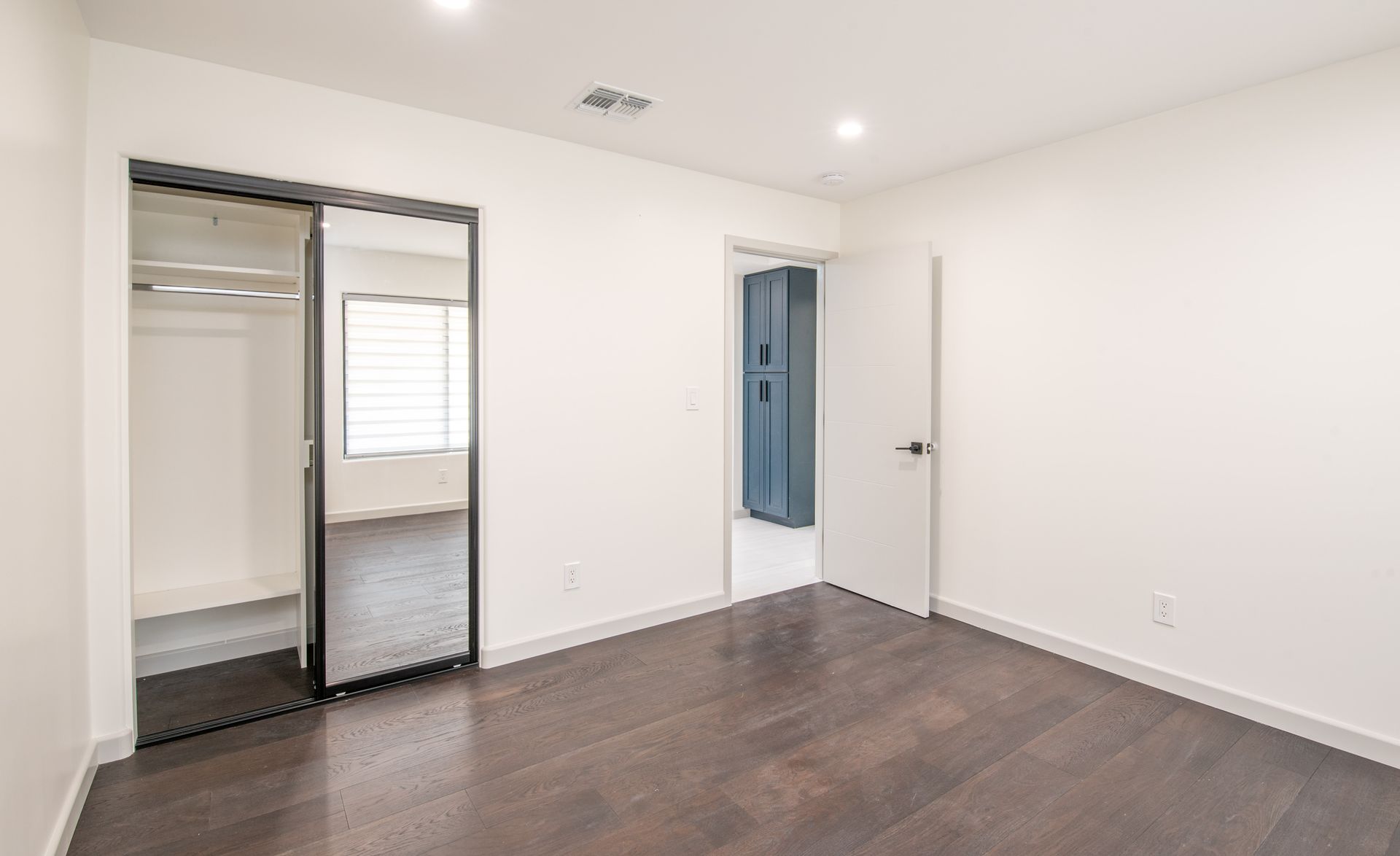 Empty bedroom with dark wood floors, a mirrored closet door, and an open door.