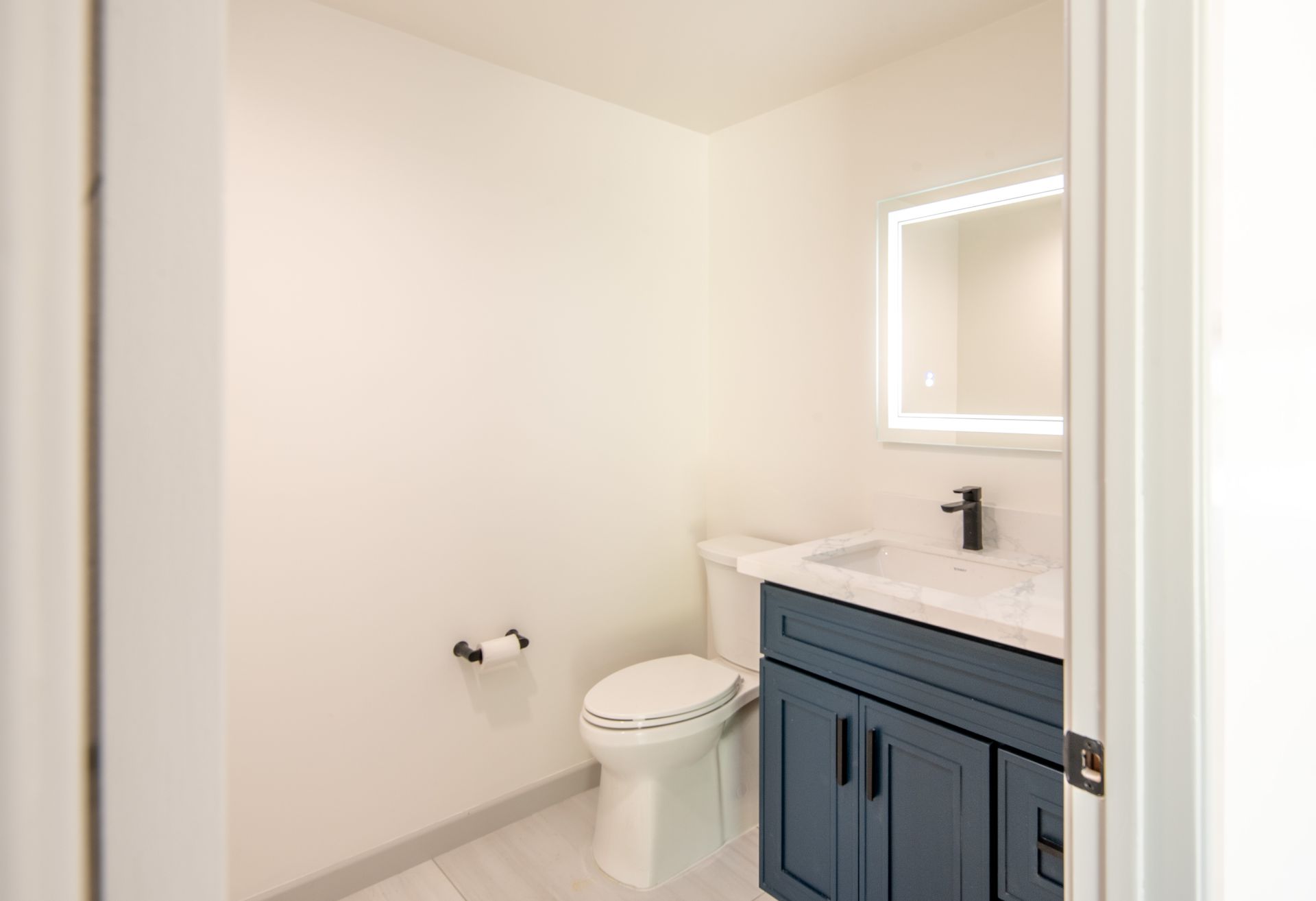 Small modern bathroom with blue vanity, white toilet, and illuminated mirror.