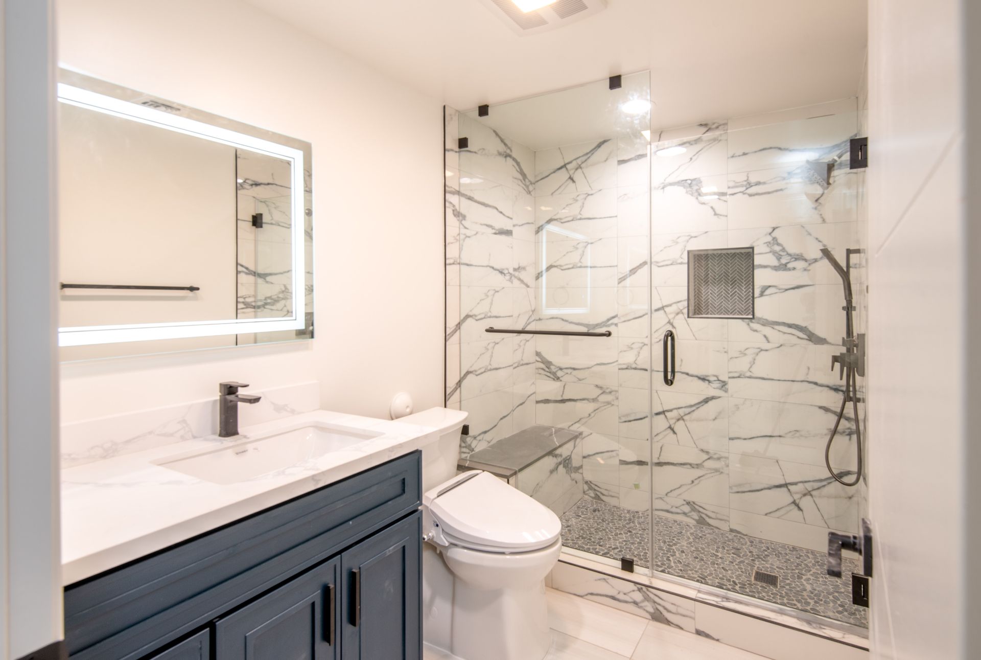 Modern bathroom with marble shower, blue vanity, illuminated mirror, and white toilet.
