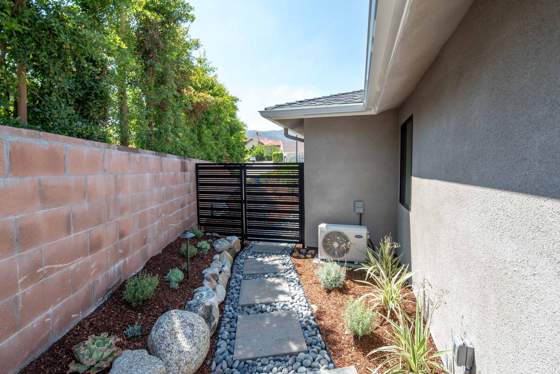 Pathway with stone pavers, gravel, and plants between a block wall and a house; black gate at end.
