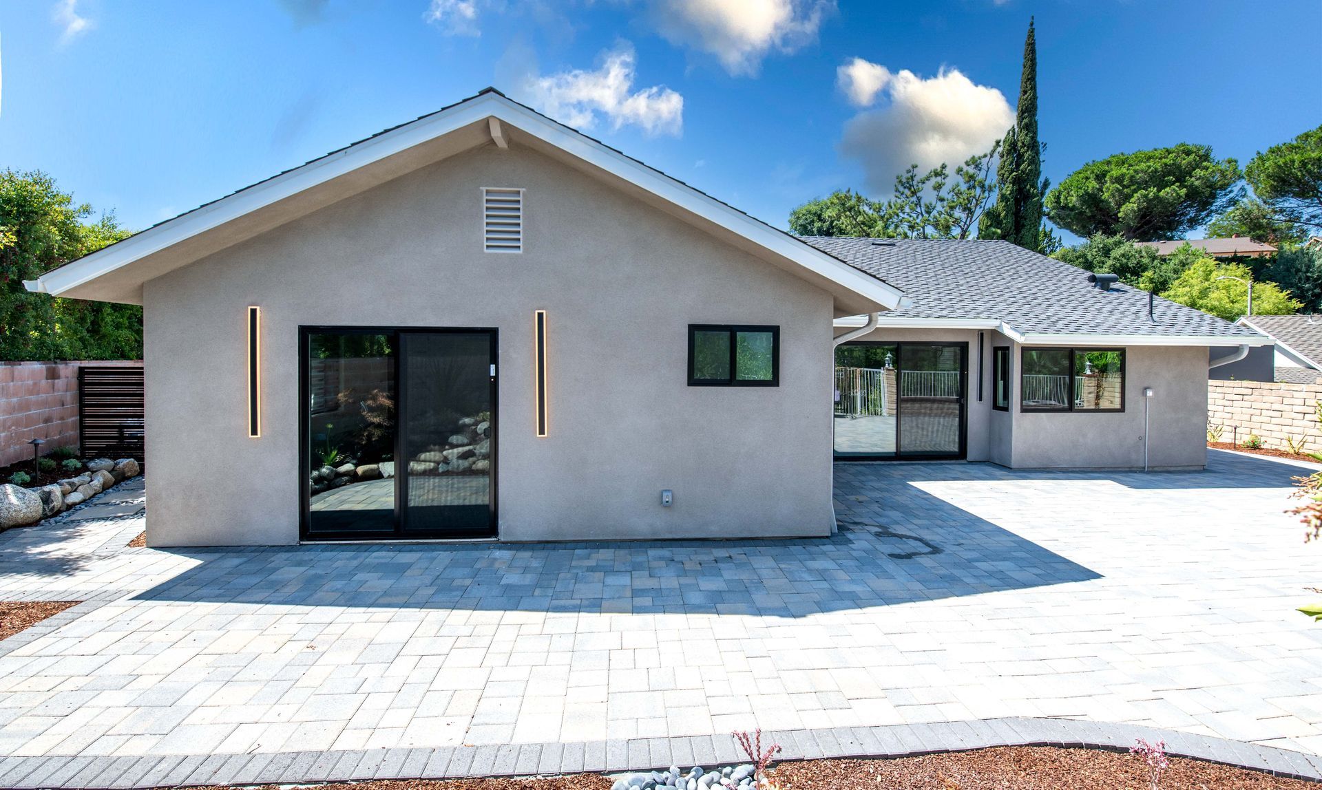 Beige single-story house with black trim, sliding doors, and a paved patio. Blue sky with trees in the background.