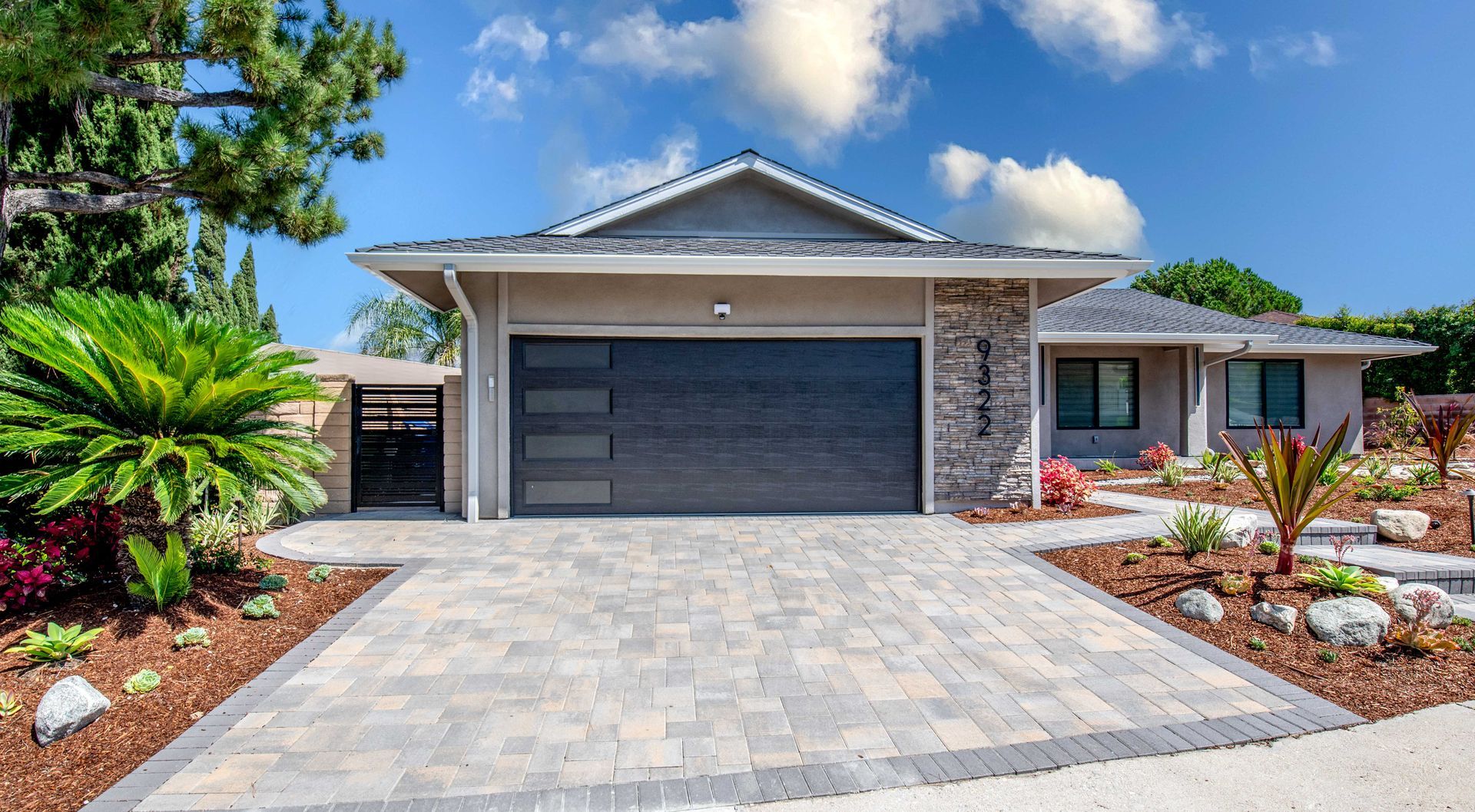 Gray house with stone accent, dark garage door, and paved driveway under a blue sky.