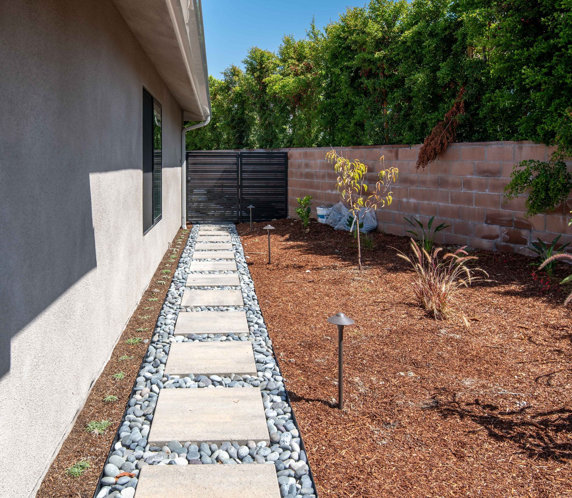 A pathway with stone pavers, black gravel, and landscaping between a stucco wall and a brown brick wall.