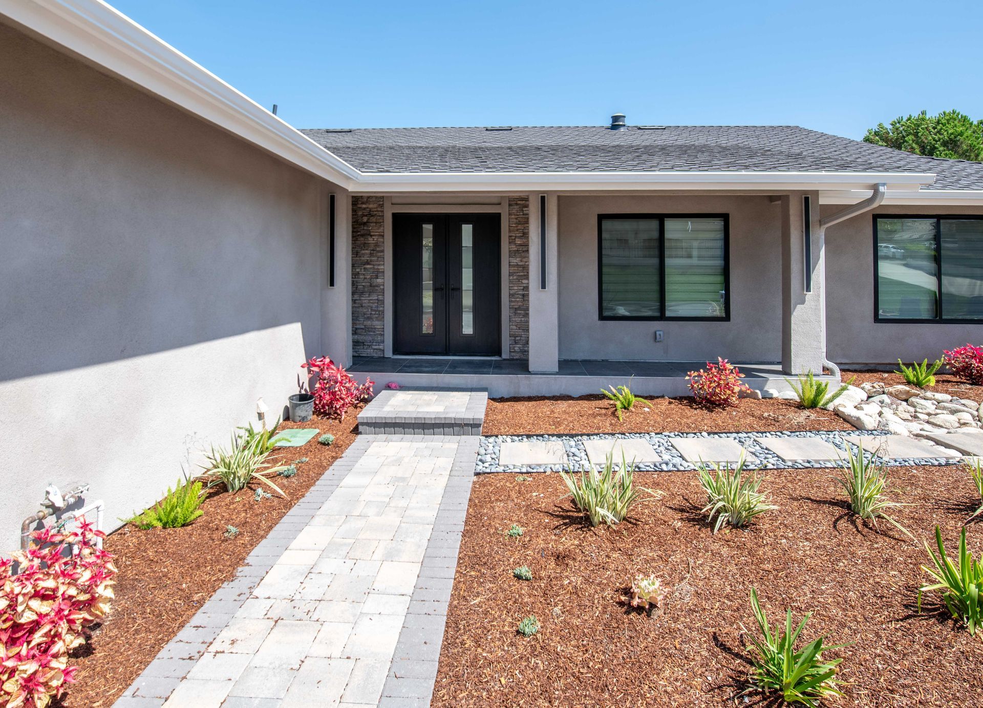 Gray house with dark door and windows, a stone walkway, and red and green plants.