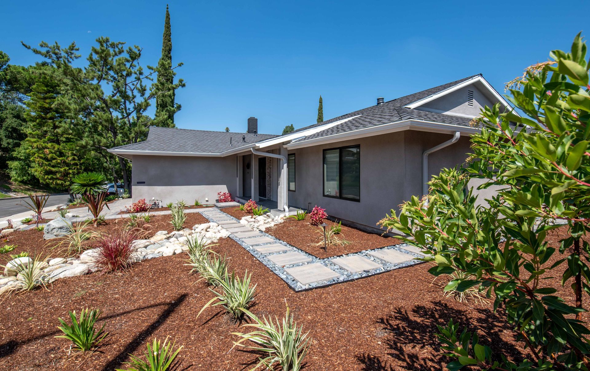 Ranch-style house with gray exterior, stone walkway, and landscaped front yard under a blue sky.