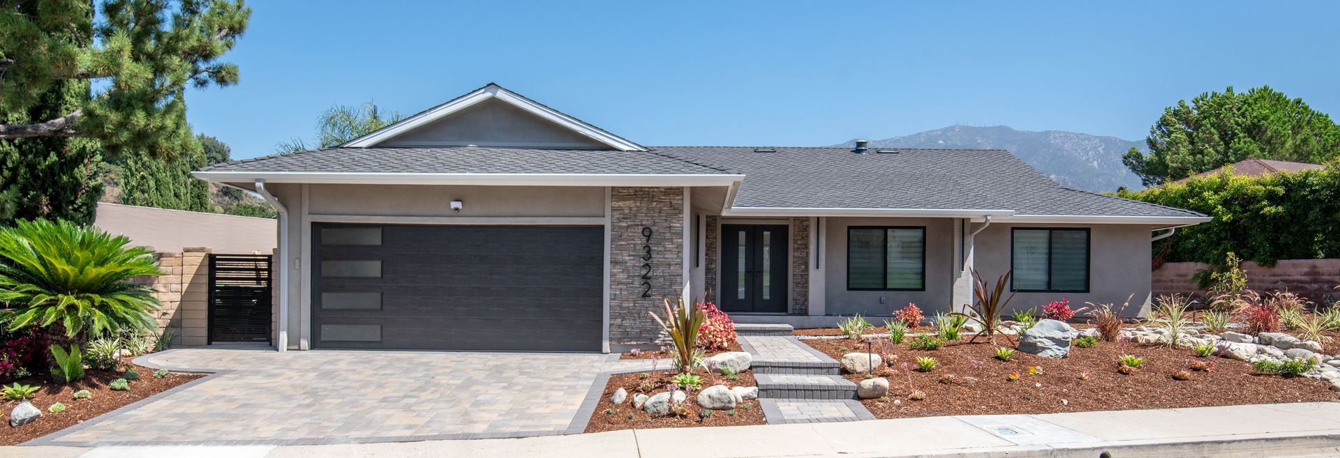 A modern, single-story house with a gray garage door, surrounded by landscaping, with mountains in the background.