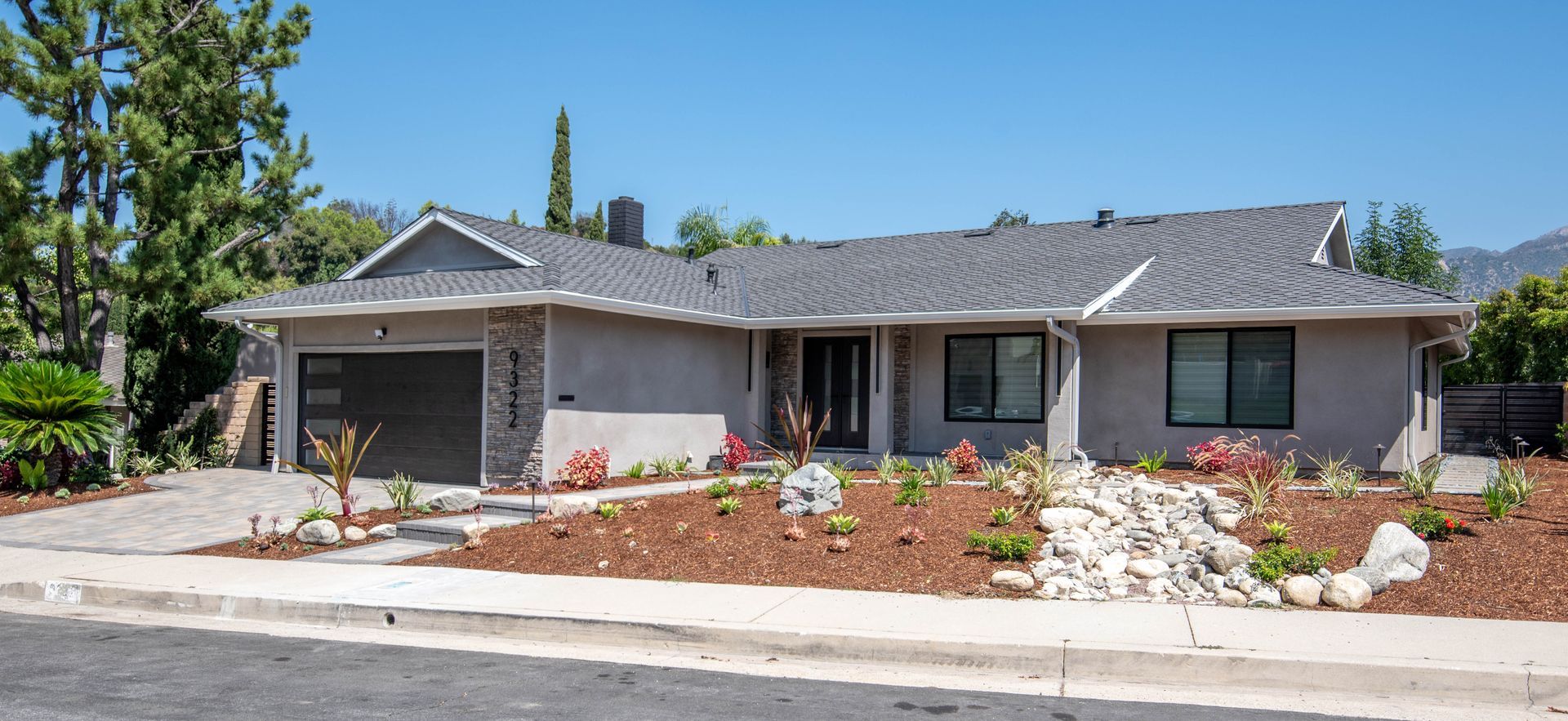 Gray house with dark roof, black garage door, landscaped yard, and blue sky.