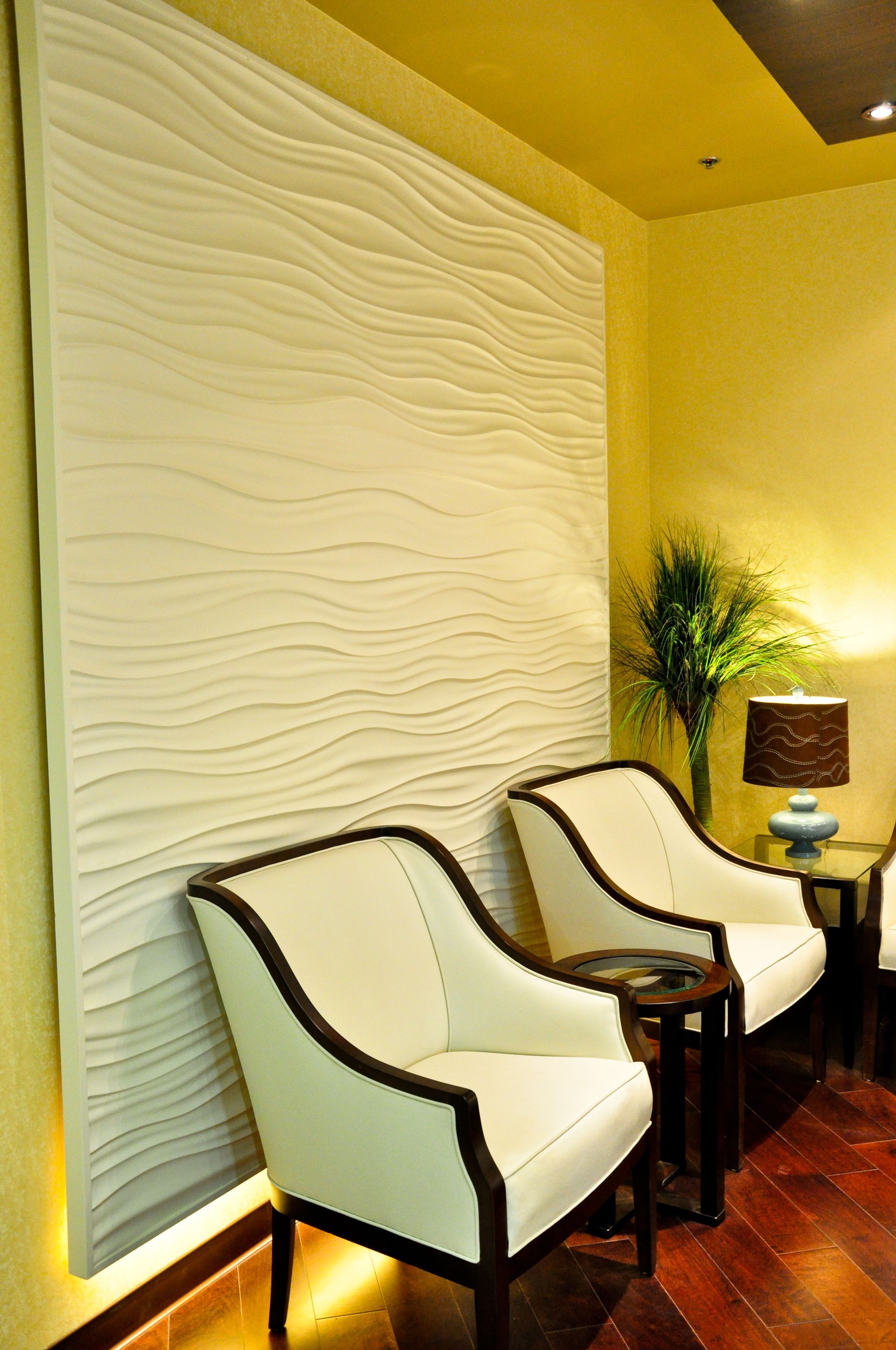 Two white chairs in front of a wave-textured wall.  Yellow walls, small table with lamp, and greenery in the corner.