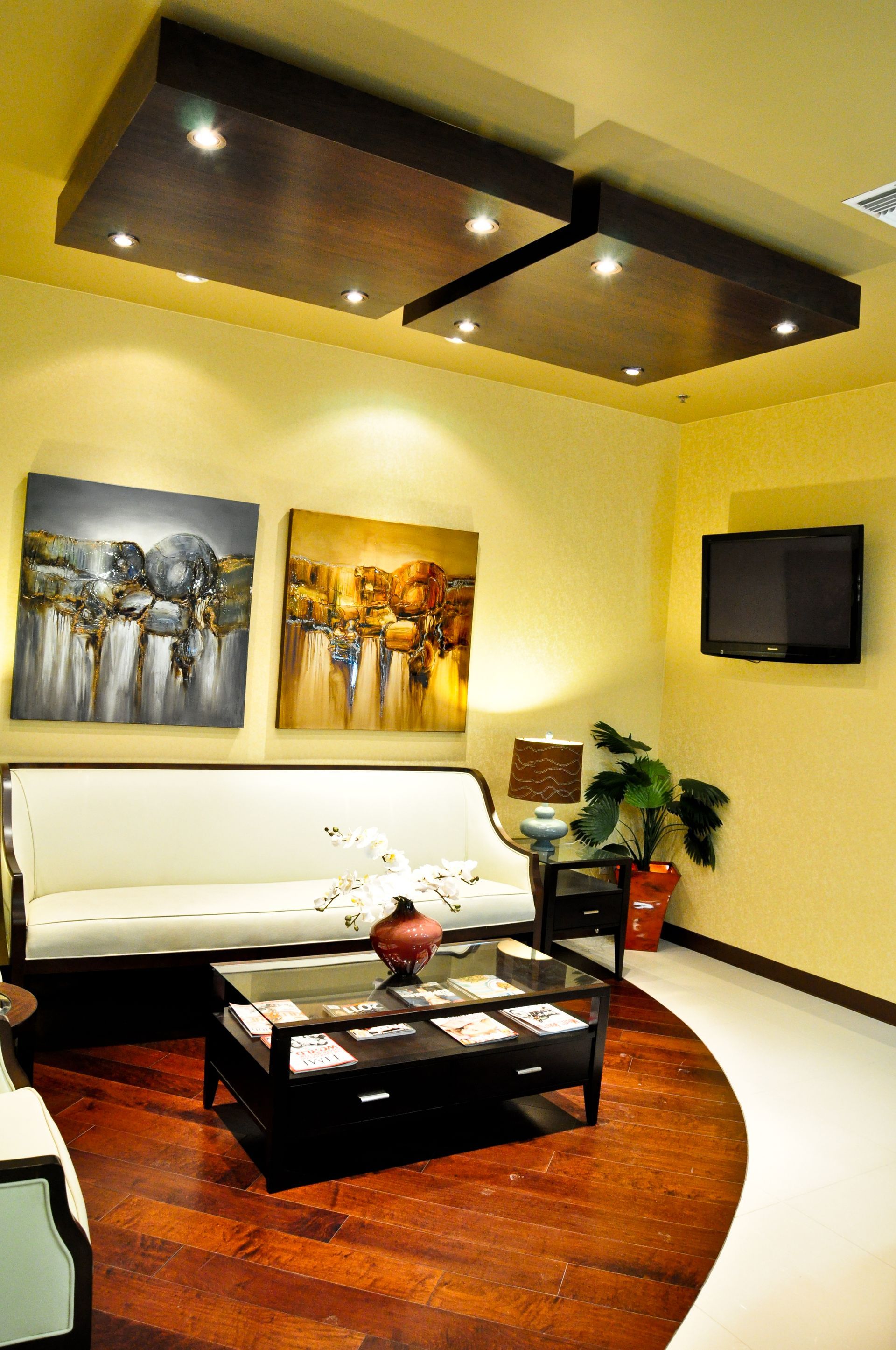 Cozy waiting room with sofa, artwork, TV, coffee table, and dark wood flooring. Ceiling has recessed lights.