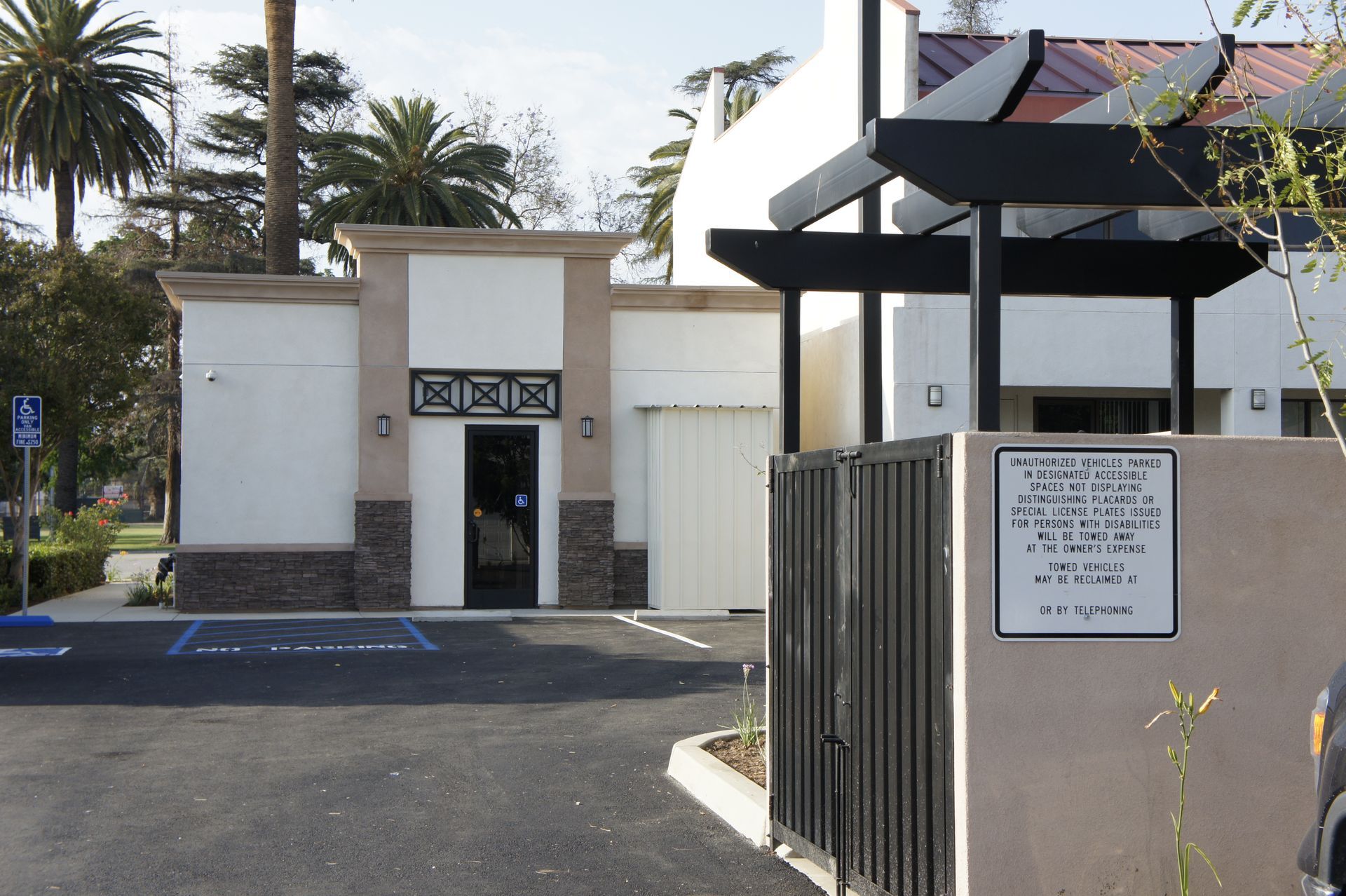 Exterior view of a low-slung building with a black fence and a sign; parking spaces and palm trees are visible.