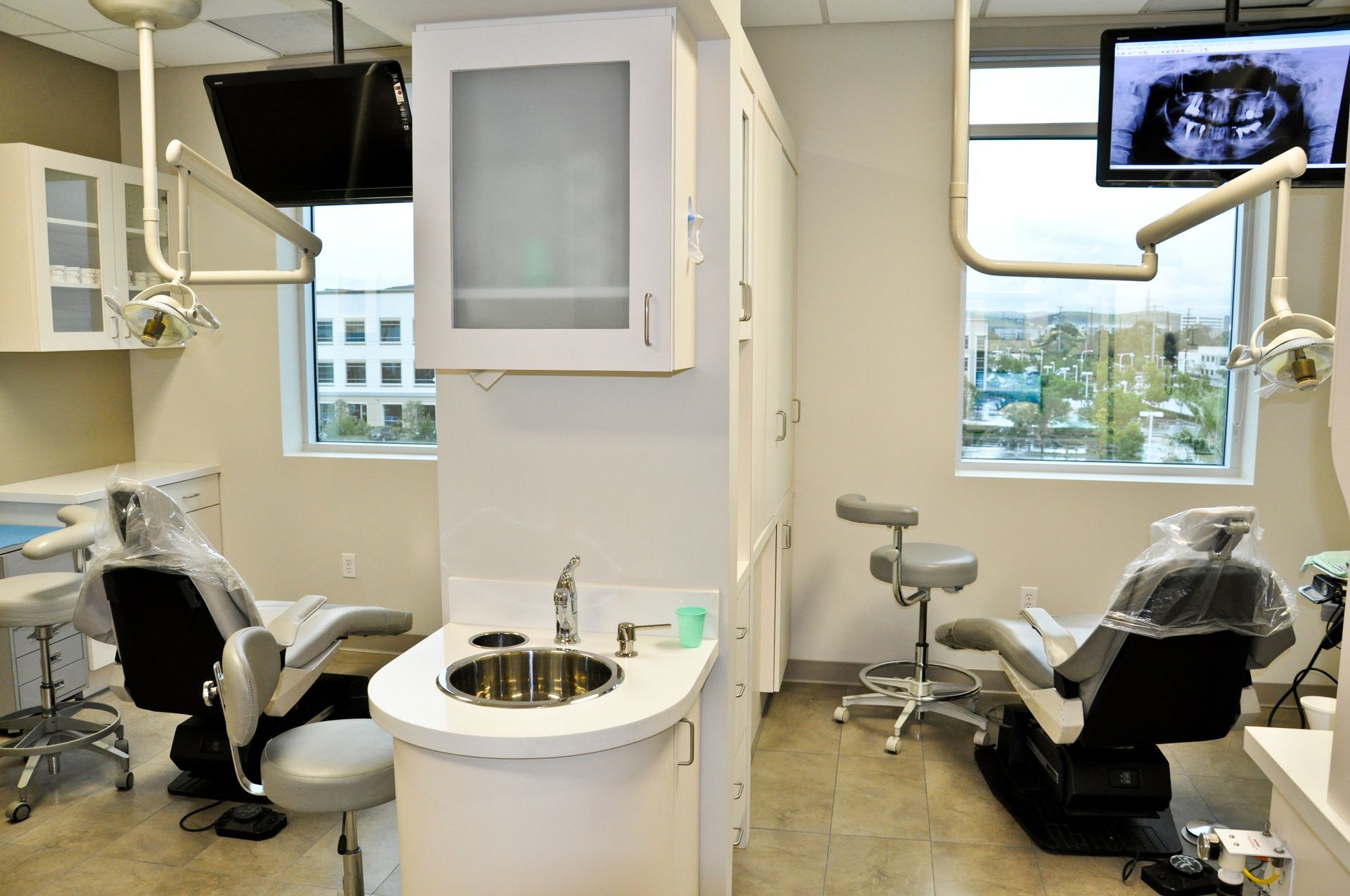 Two dental chairs in an office. Cabinets, sink, and monitors are visible. Daylight streams through windows.