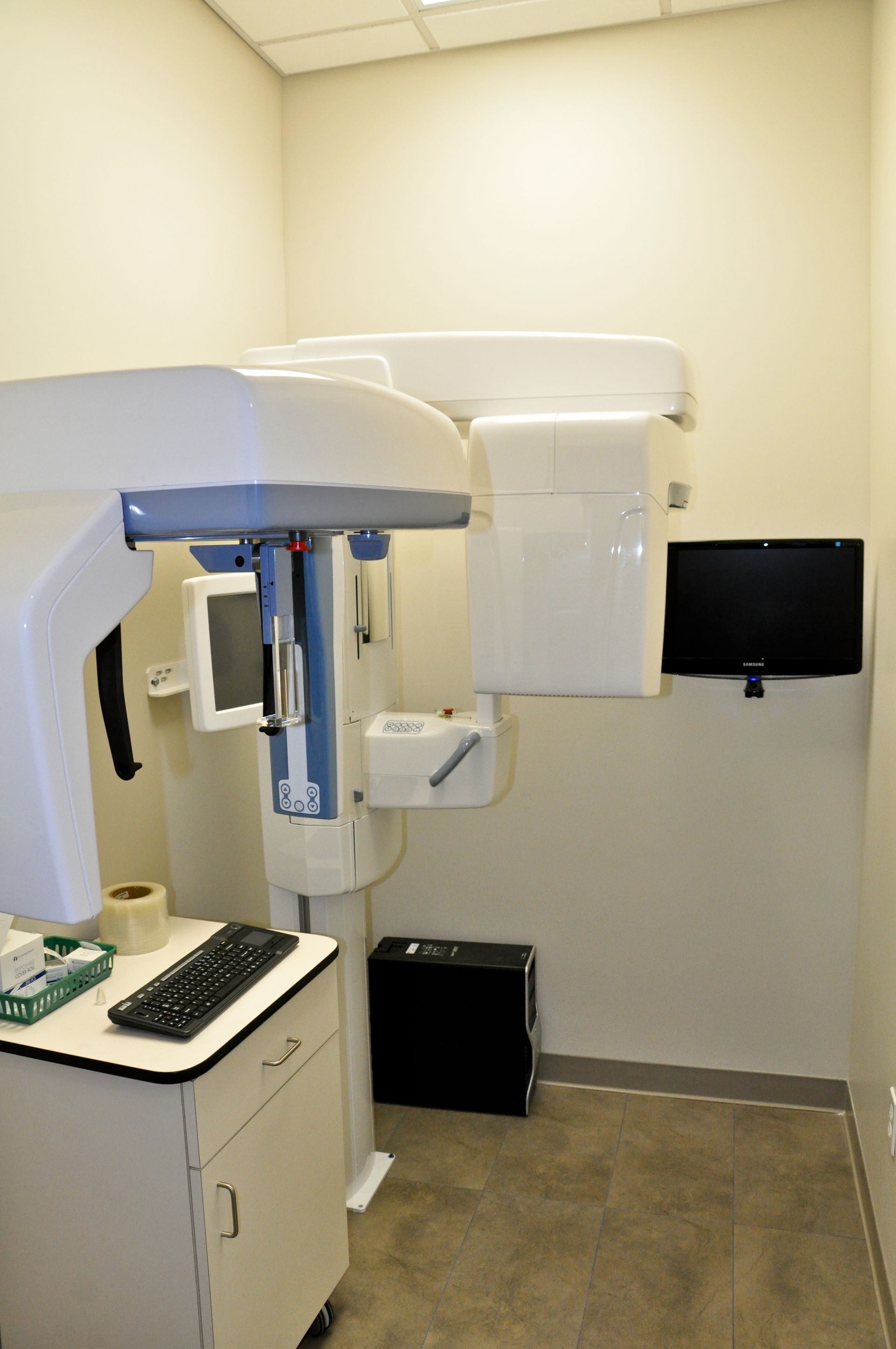 Dental X-ray machine in a dentist's office. White equipment, a computer screen on the wall, and a small cabinet.