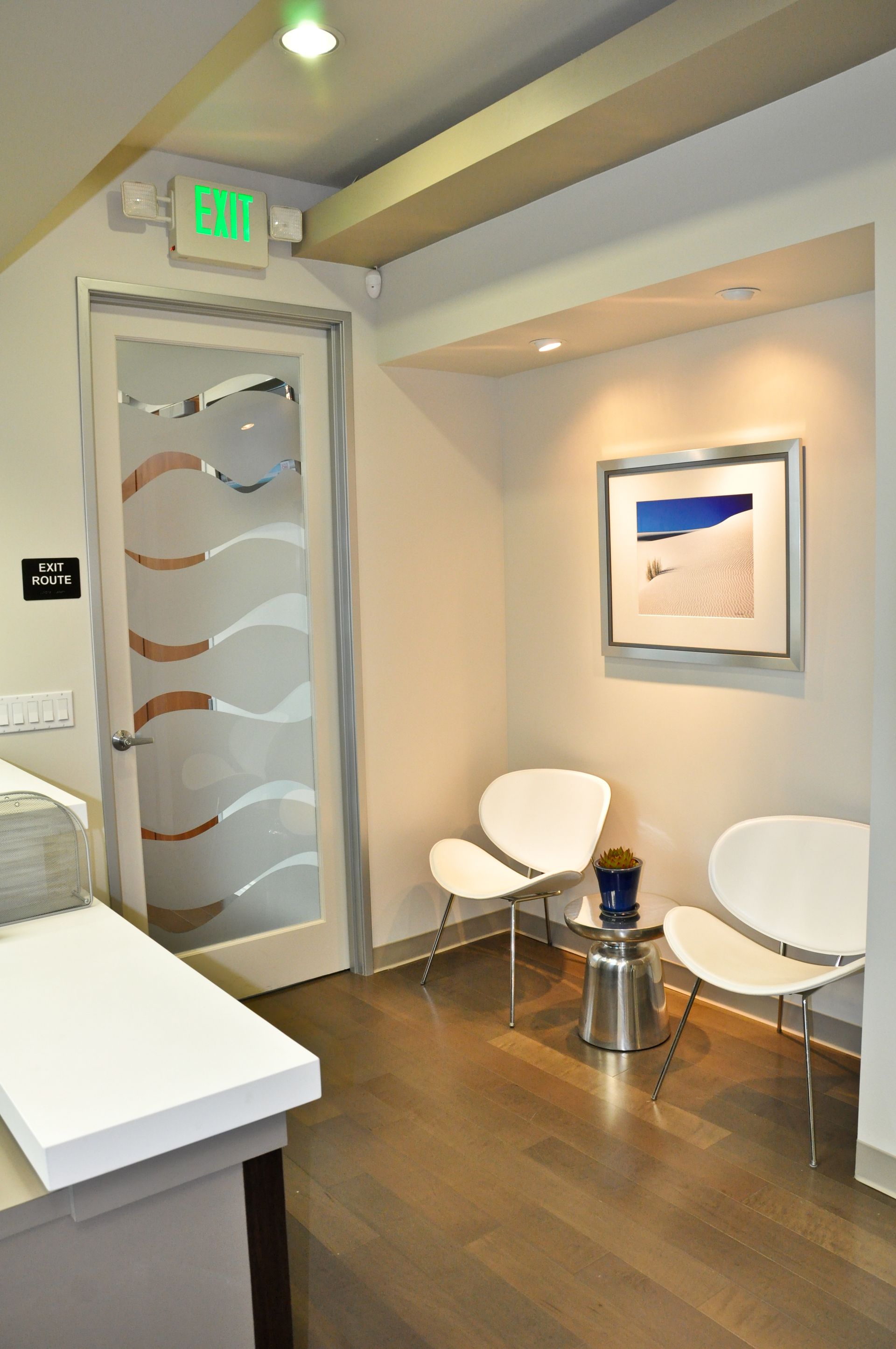 Waiting room with white chairs, framed art, frosted glass door, and dark wood floor.