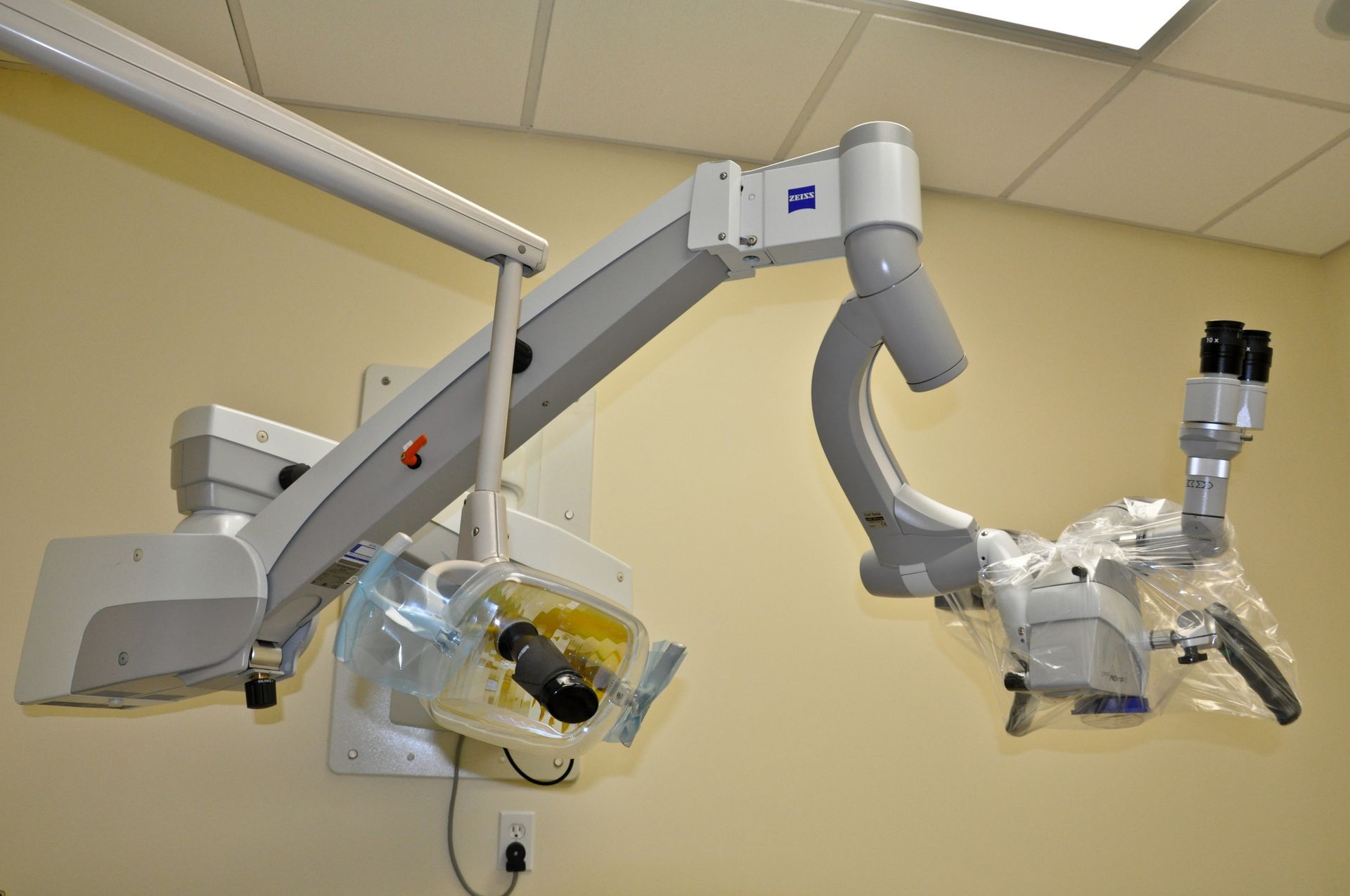 Medical operating microscope mounted on a wall. Gray and white with attached clear plastic.