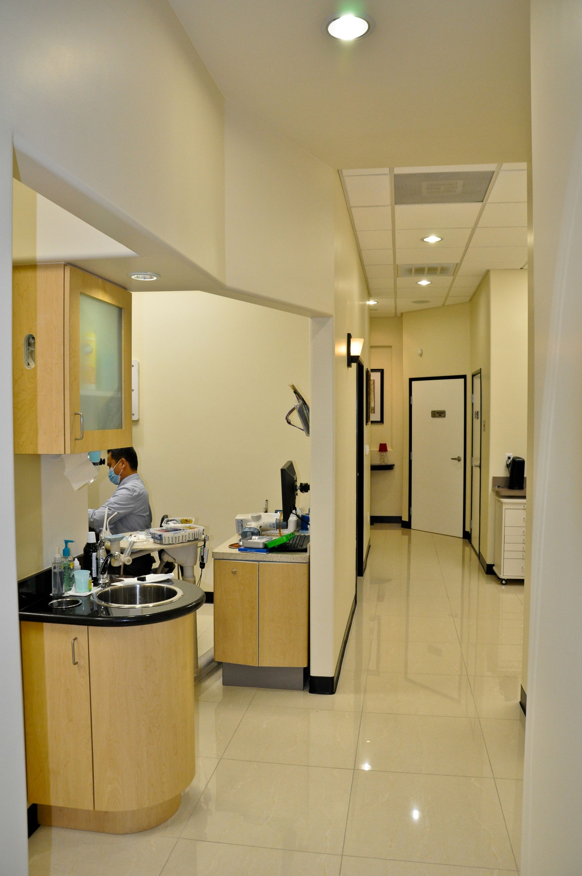 Long hallway in a dentist's office with tan walls and floors, leading to white doors. A dentist is working in the foreground.