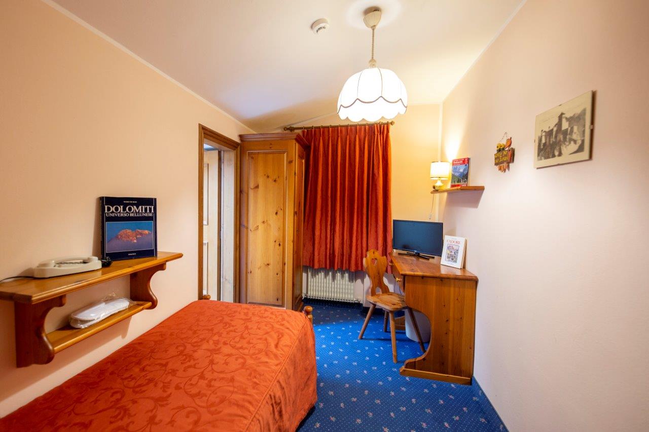 Piccola camera da letto con copriletto rosso, tappeto blu, scrivania e tende arancioni.