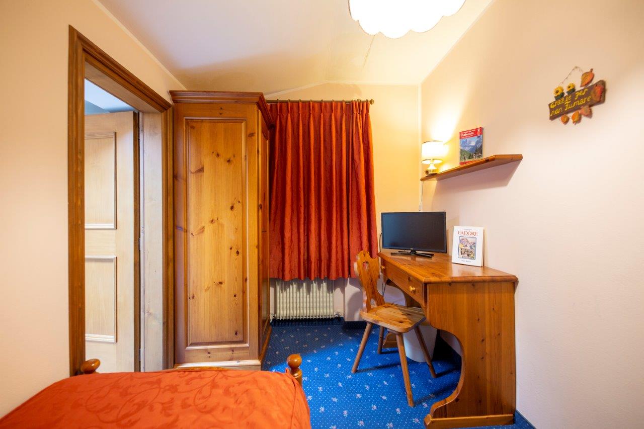 Piccola camera d'albergo con mobili in legno, tende rosse, una scrivania con TV e un tappeto blu.