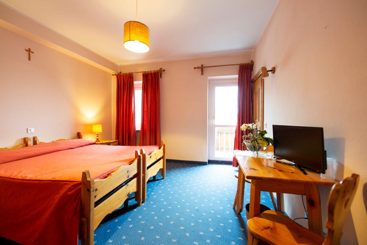 Luminosa camera d'albergo con due letti singoli, moquette blu, tende rosse e un piccolo televisore su un tavolino di legno.