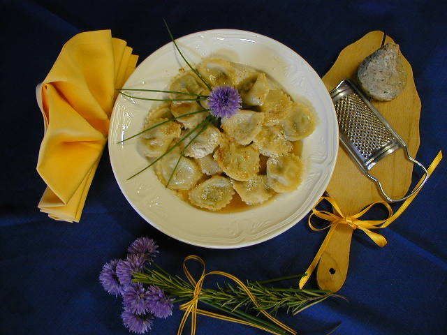 Tovagliolo e cucchiaio gialli accanto a un piatto di ravioli su un tavolo blu, con fiori viola come guarnizione.