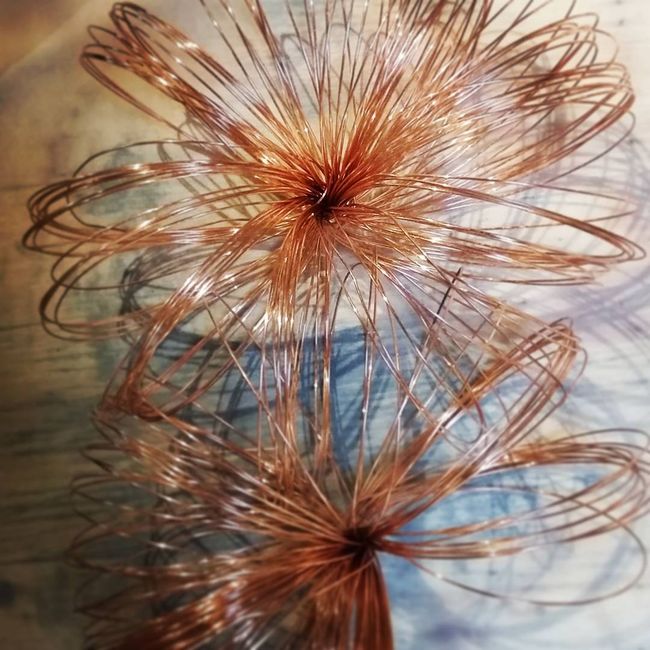 Scultura floreale in filo di rame con due fiori circolari e appuntiti su uno sfondo sfocato.