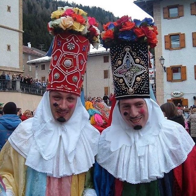 Due figure in costume con alti cappelli decorati e gorgiere bianche a una festosa parata all'aperto.
