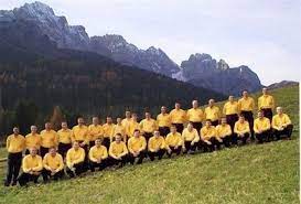 Un folto gruppo di persone con indosso magliette gialle posa su un pendio erboso con le montagne sullo sfondo.