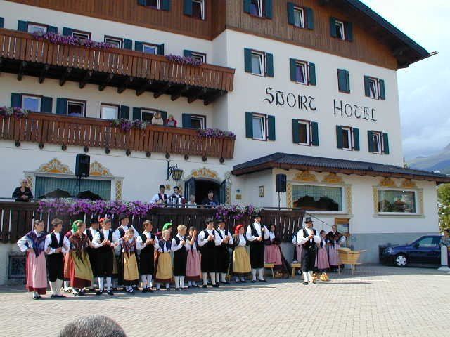 Gruppo di persone in abiti tradizionali in posa davanti a un hotel con le montagne sullo sfondo.