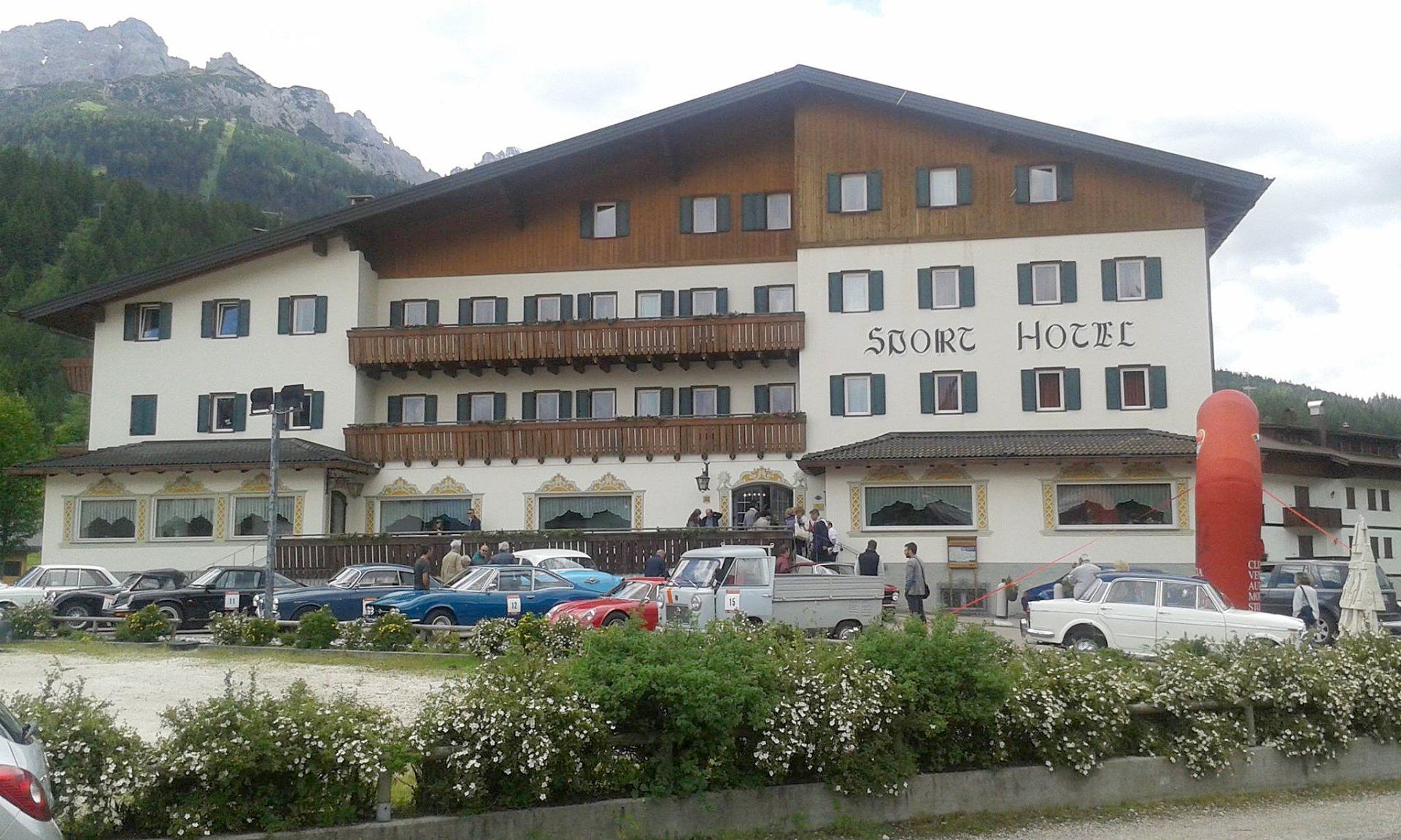 Ampio hotel alpino con facciata bianca e marrone, auto parcheggiate e montagne sullo sfondo.