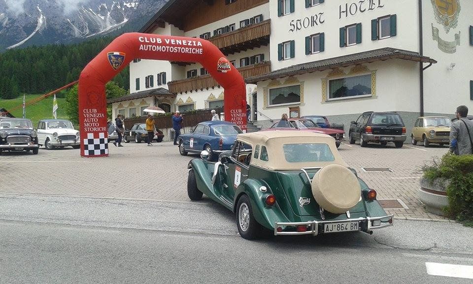 Una decappottabile verde d'epoca parcheggiata fuori da un hotel, sotto un arco rosso allestito per eventi, con le montagne sullo sfondo.